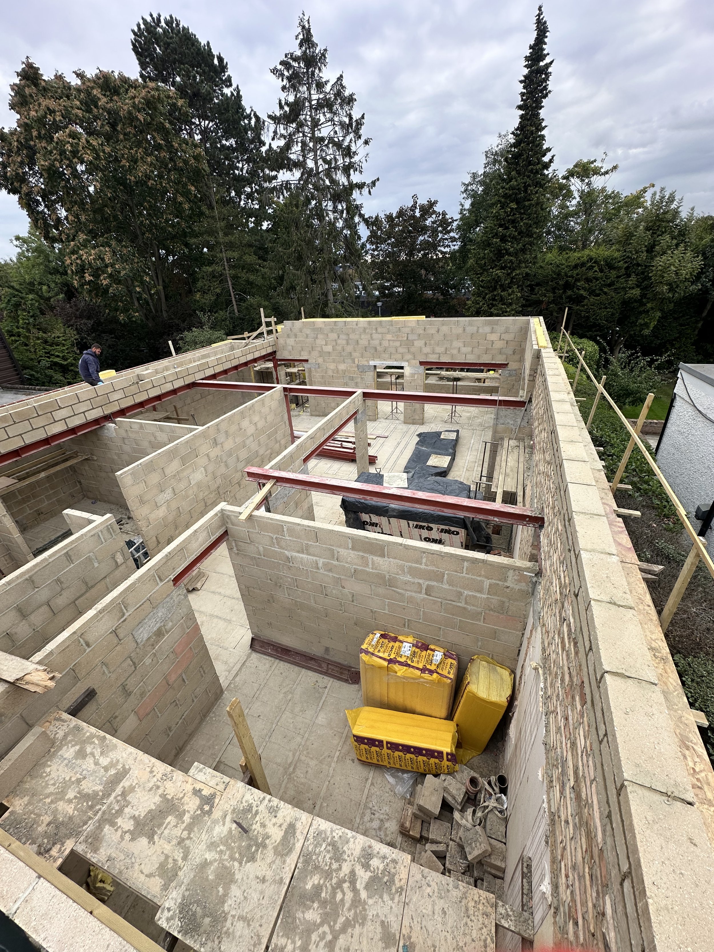 Construction site of a building with brick walls under development, construction materials, and equipment visible, surrounded by trees.