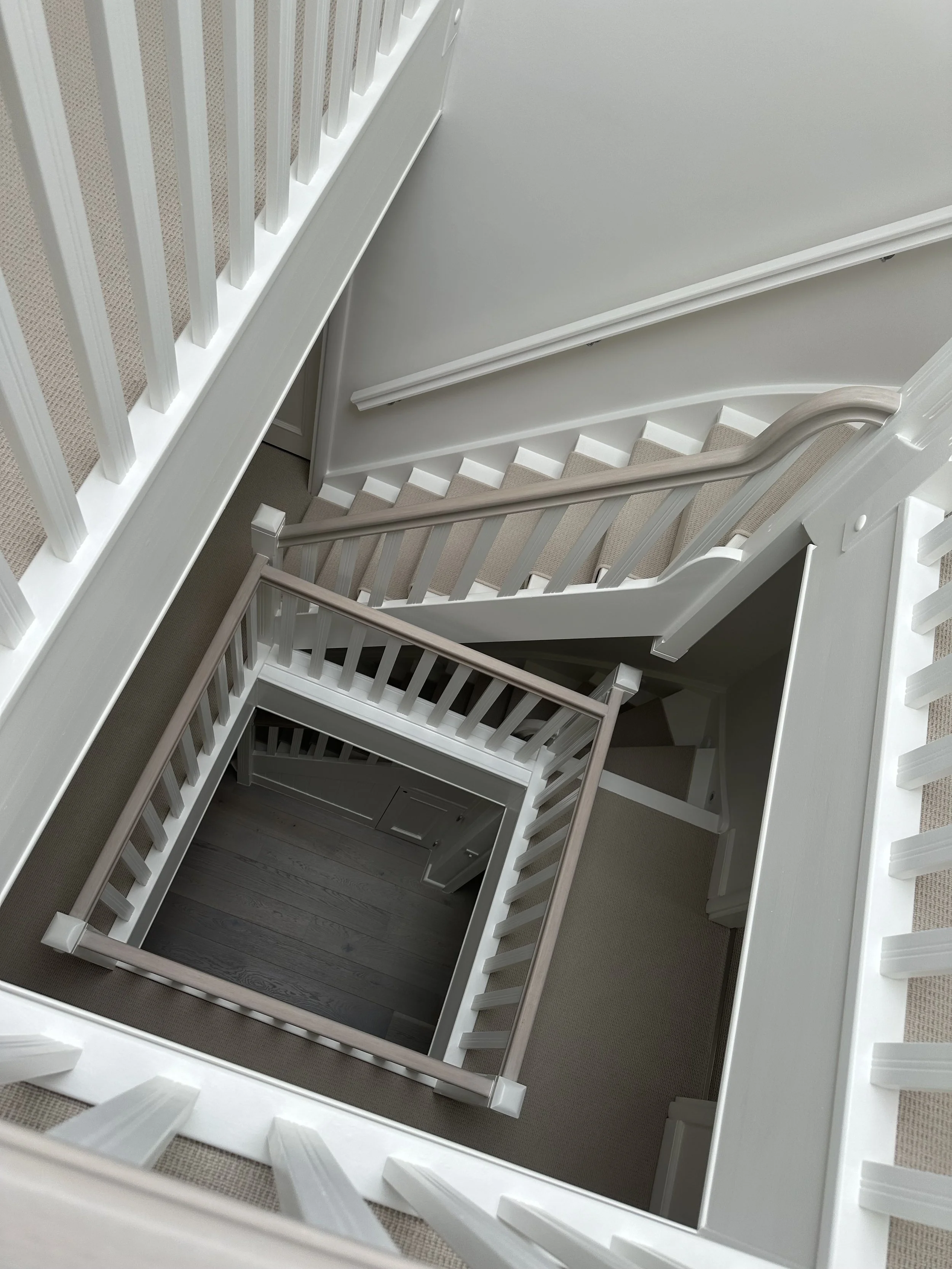 Top-down view of a square staircase with beige carpeted steps and white railings, looking down to the lower floors.