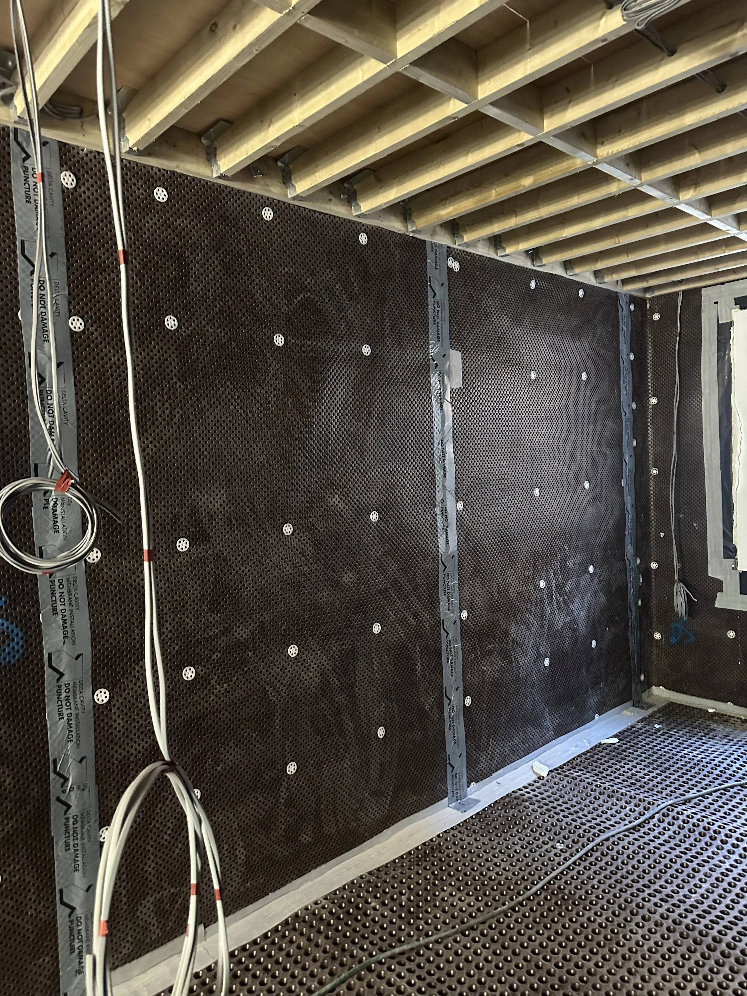 Insulated wall with subfloor, exposed ceiling with wooden beams, and electrical and plumbing wiring in a construction or renovation site.