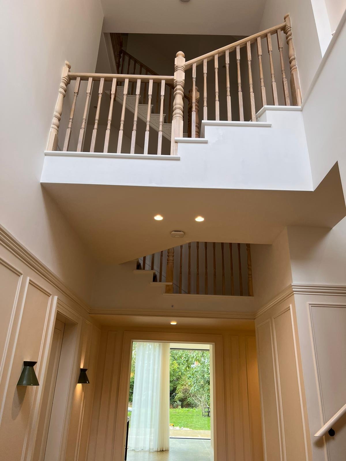 Interior view of a multi-story house with wooden railings, staircase, and large window with sheer curtains overlooking a garden.