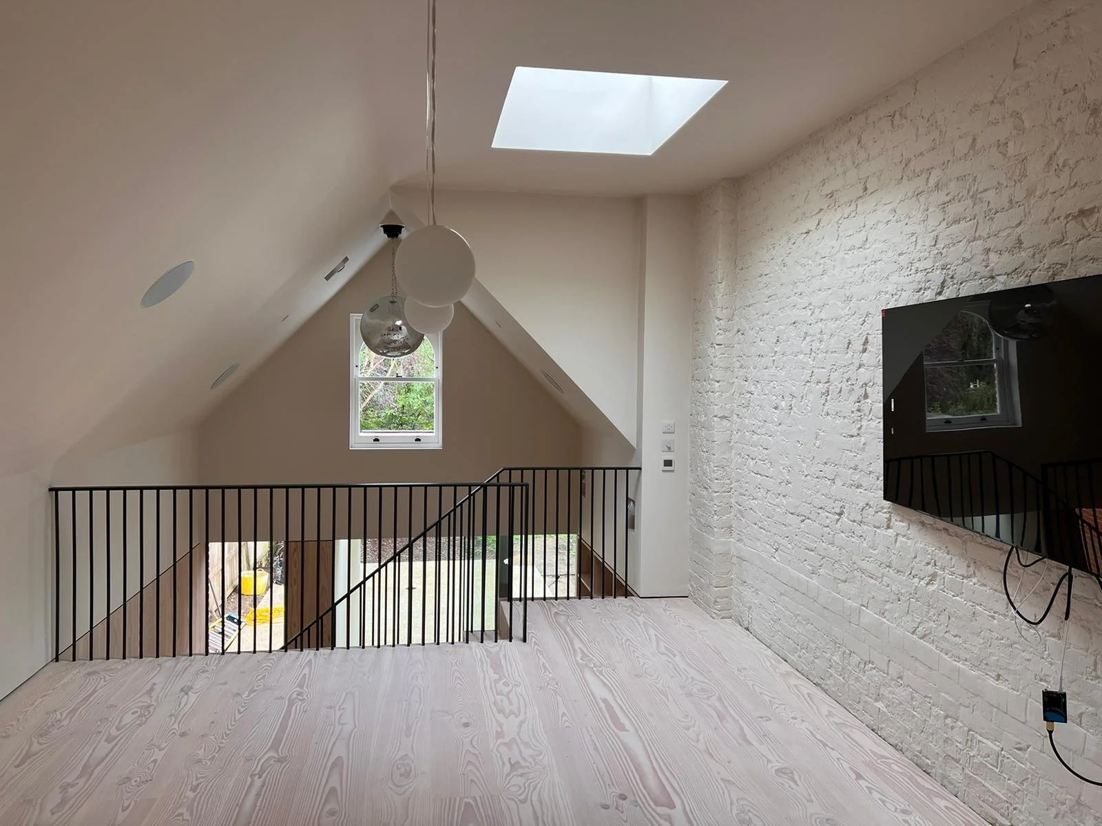 Interior view of an attic space with sloped ceilings, a skylight, a small window, a white brick accent wall, a mounted TV, a black railing, and decorative hanging lights.