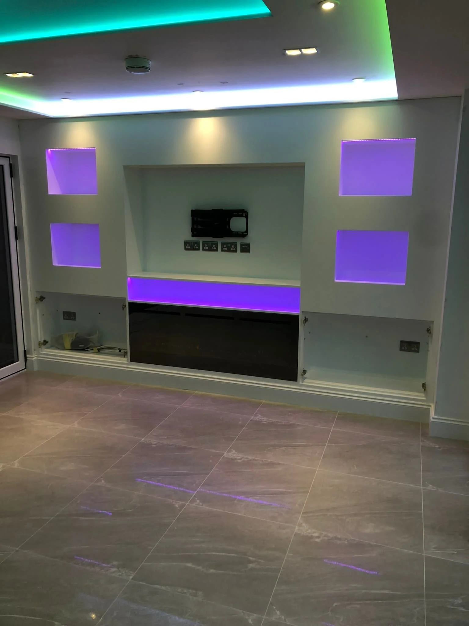 Empty built-in entertainment center with purple and blue LED lighting and wall outlets, in a modern living room with gray tiled floor.