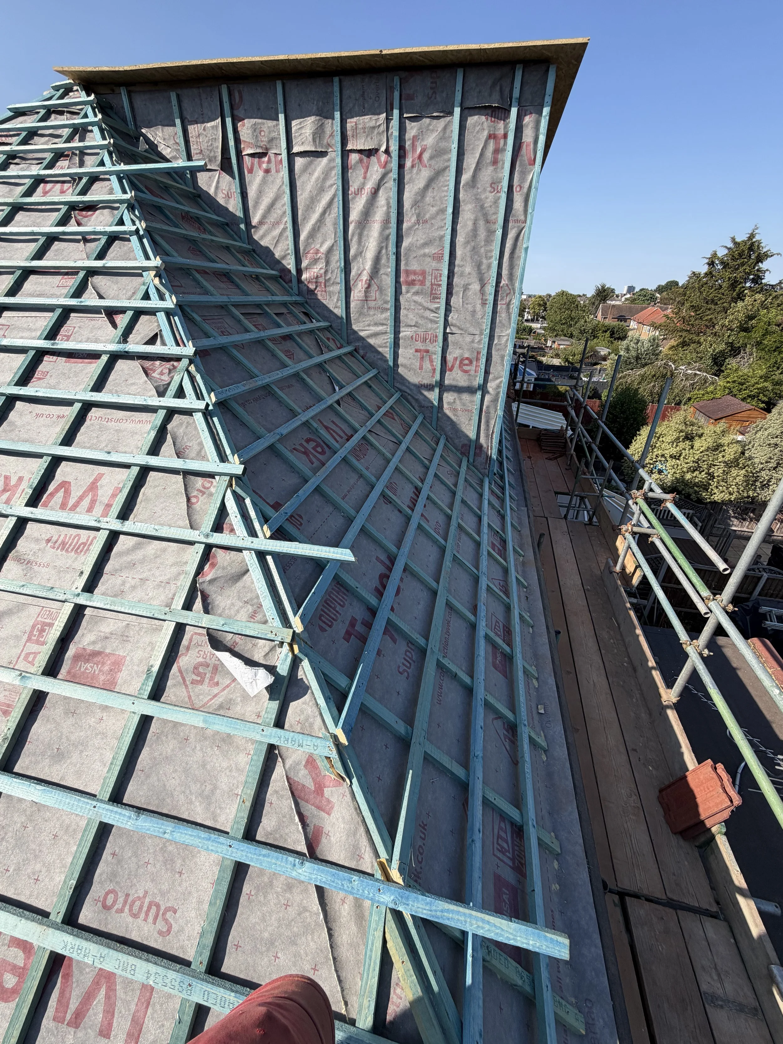 Roof under construction with wooden framing and insulation, scaffolding around the edges, and residential houses with trees and a clear sky in the background.