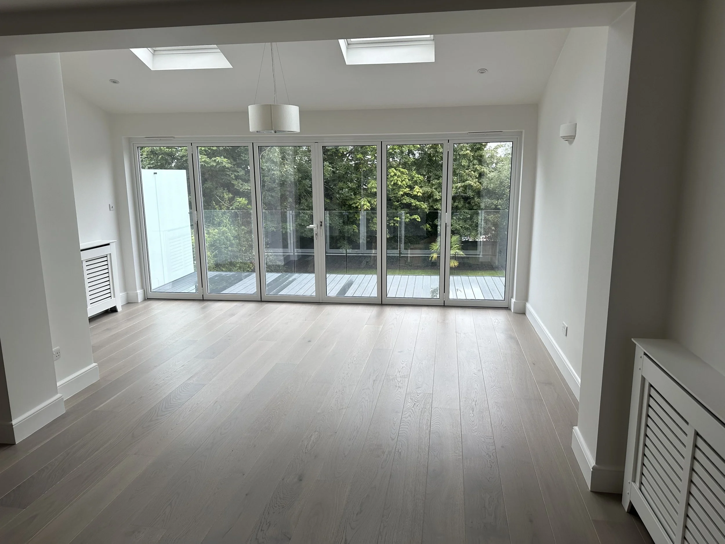 Empty modern room with light wood flooring, white walls, and large glass sliding doors leading to an outdoor balcony with trees visible outside.
