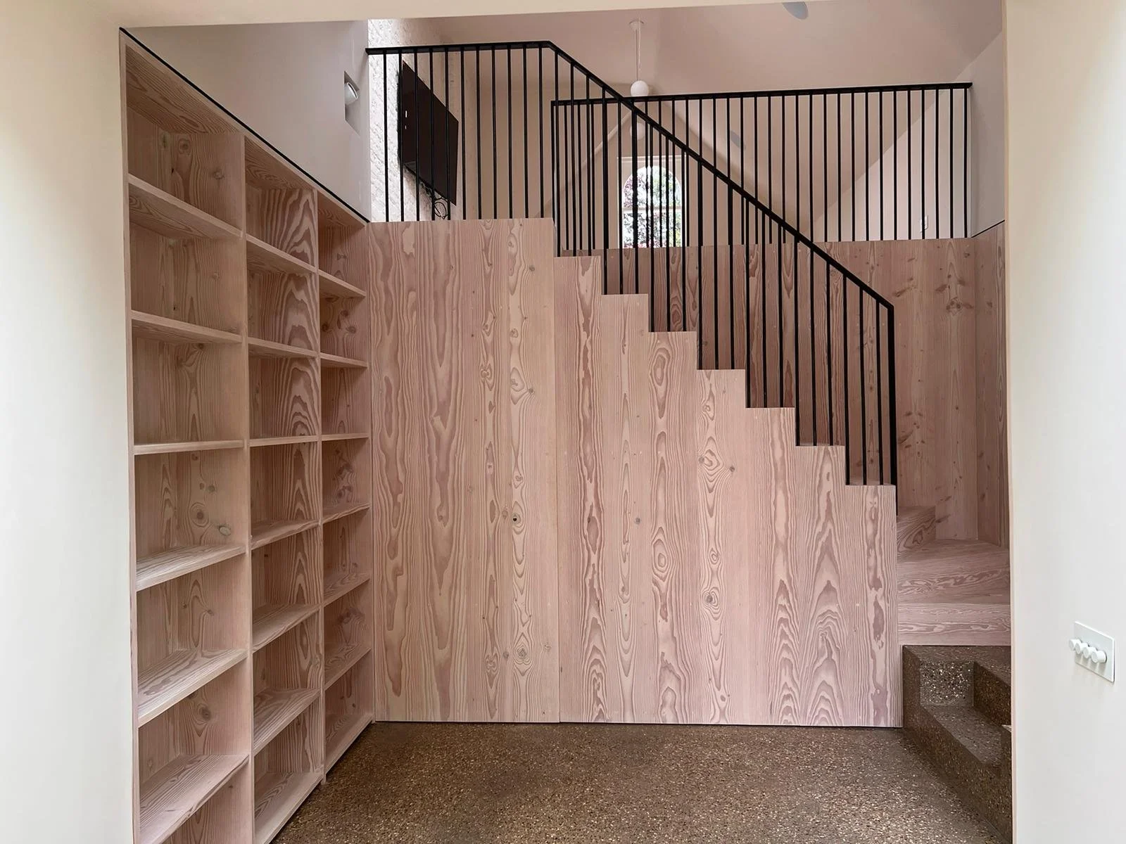 Interior staircase with a black metal railing, light wood paneling, empty shelves on the left, and a small step at the bottom right.