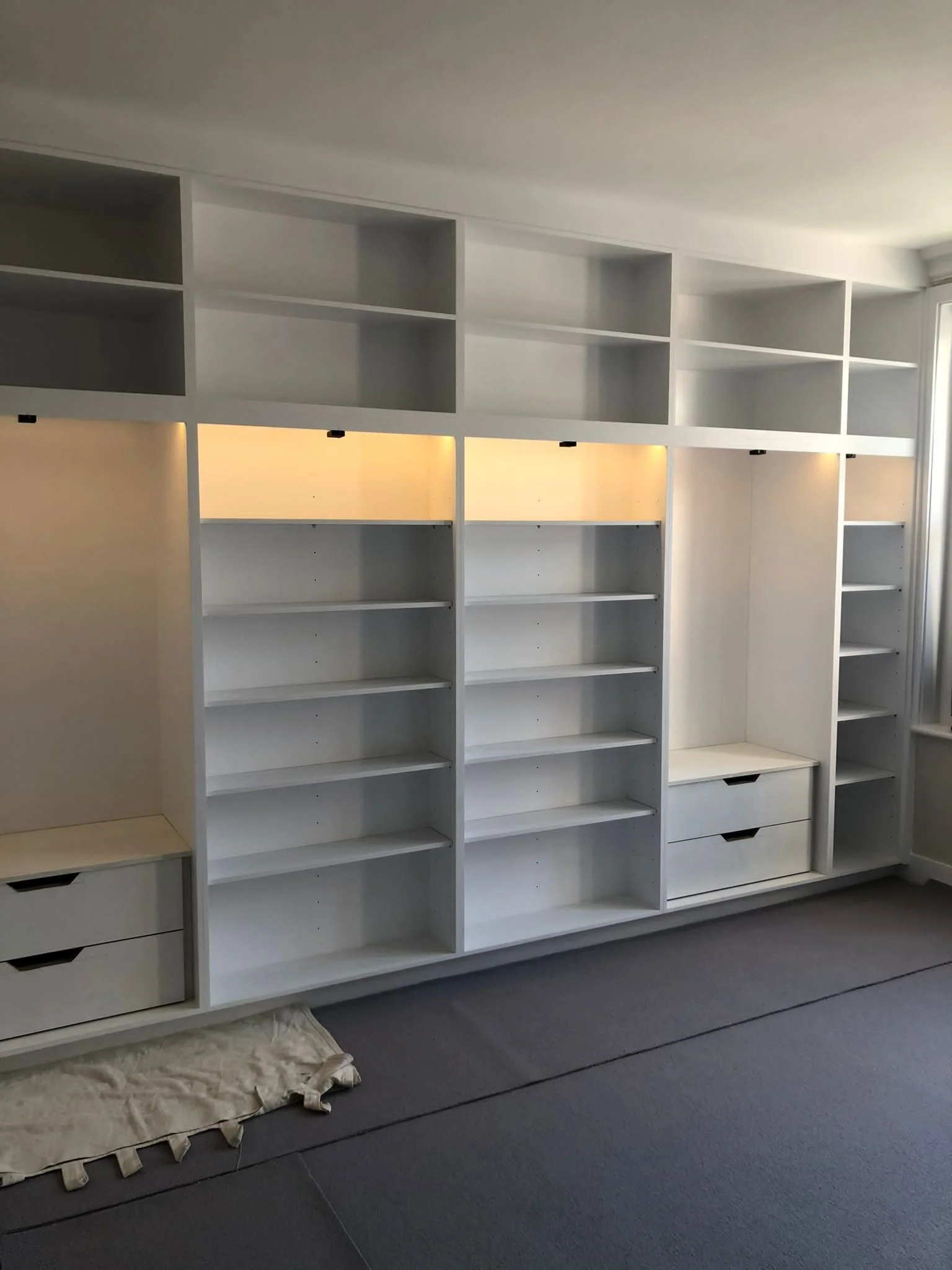 Empty white built-in shelves and drawers in a room with gray carpet and a window.