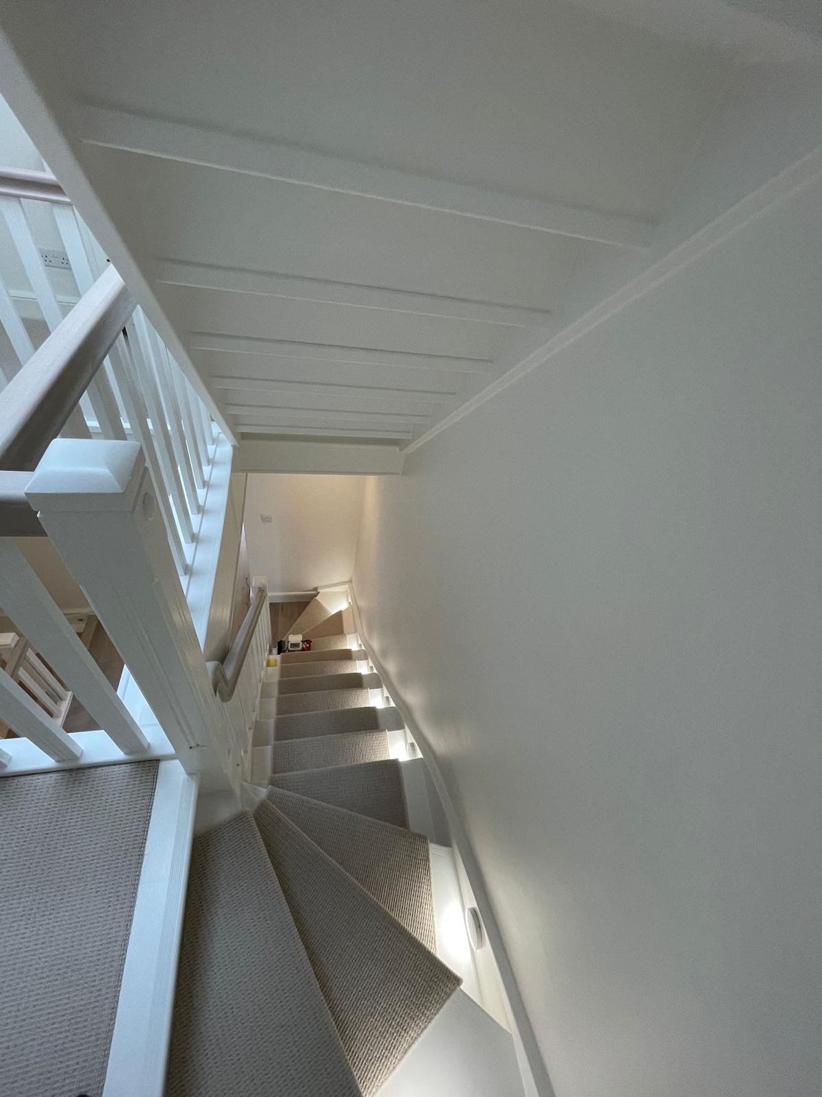 Indoor view of a staircase with gray carpeting, white walls, a curved wall at the bottom, and a white railing with brown handrail on the left side.