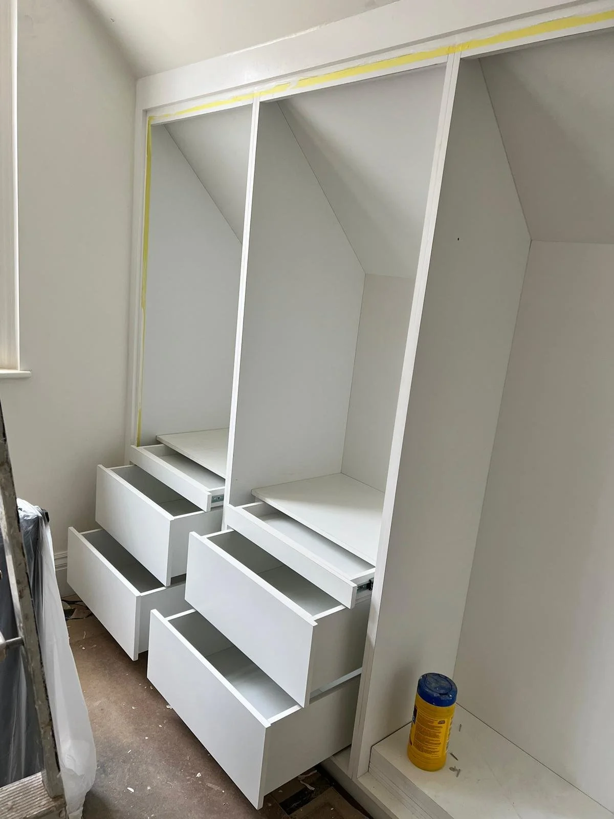  white built-in closet with drawers and shelves under construction in a room, with a can of paint or wood finish on the floor.