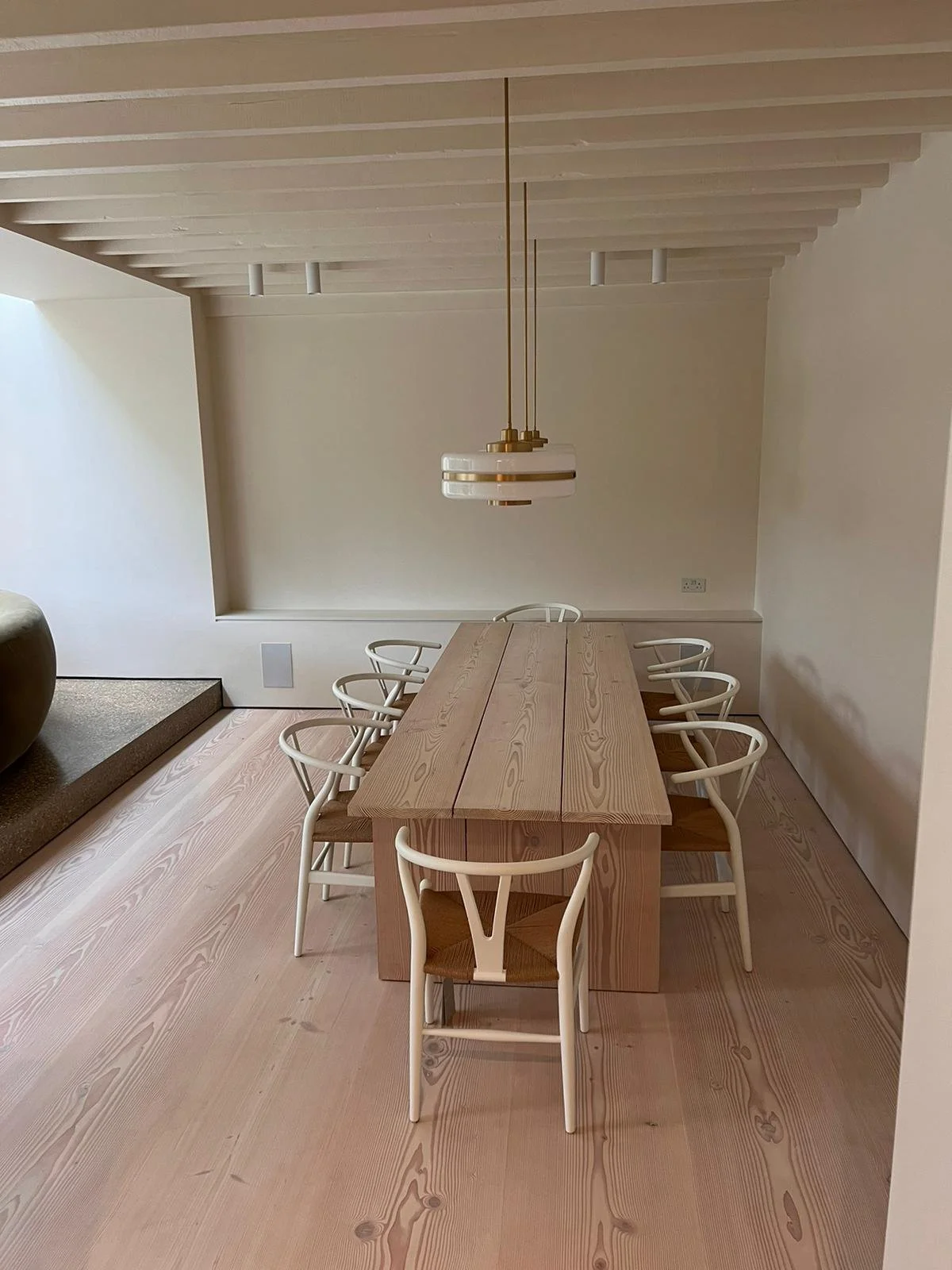 A minimalist dining room with a rectangular wooden table, surrounded by eight white chairs with woven seats, and a modern chandelier hanging from the ceiling.