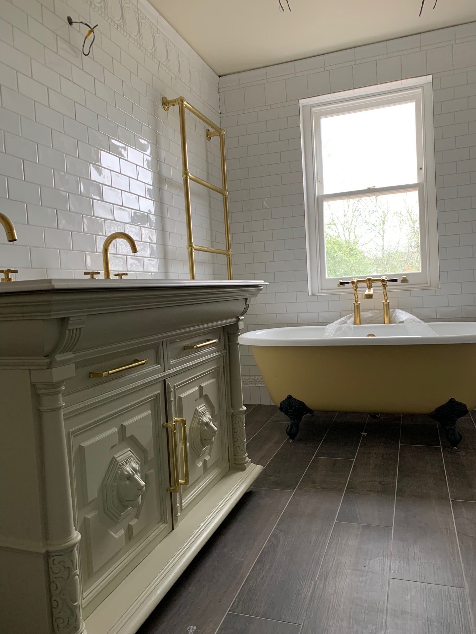 A bathroom with a white tiled wall, a vintage-style cream vanity with lion head accents, and a clawfoot bathtub with gold fixtures, located next to a window that shows outside greenery.