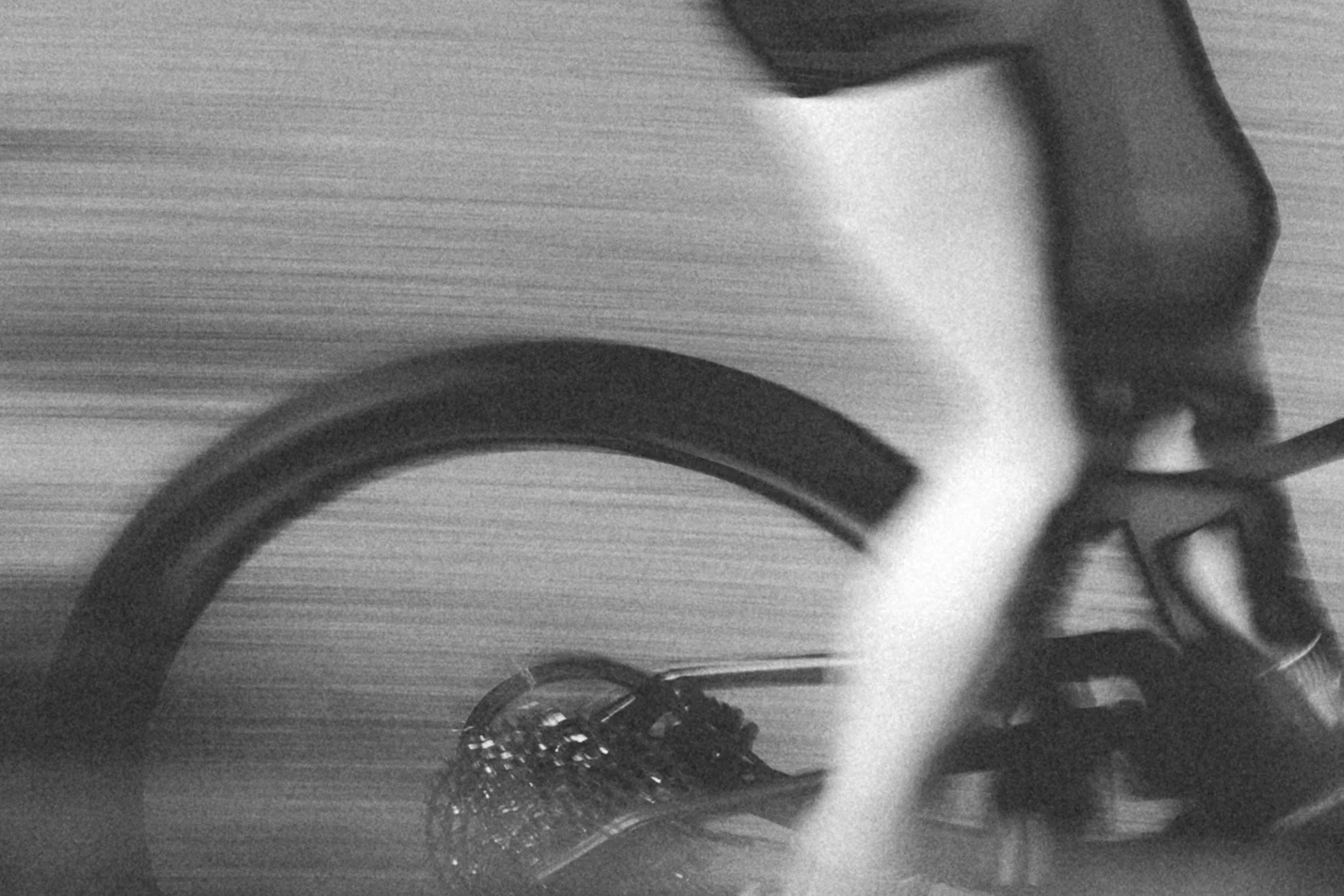 Une photo en noir et blanc d'un vélo avec le cadre, la roue avant et la pédale visibles, posé sur un sol en bois.