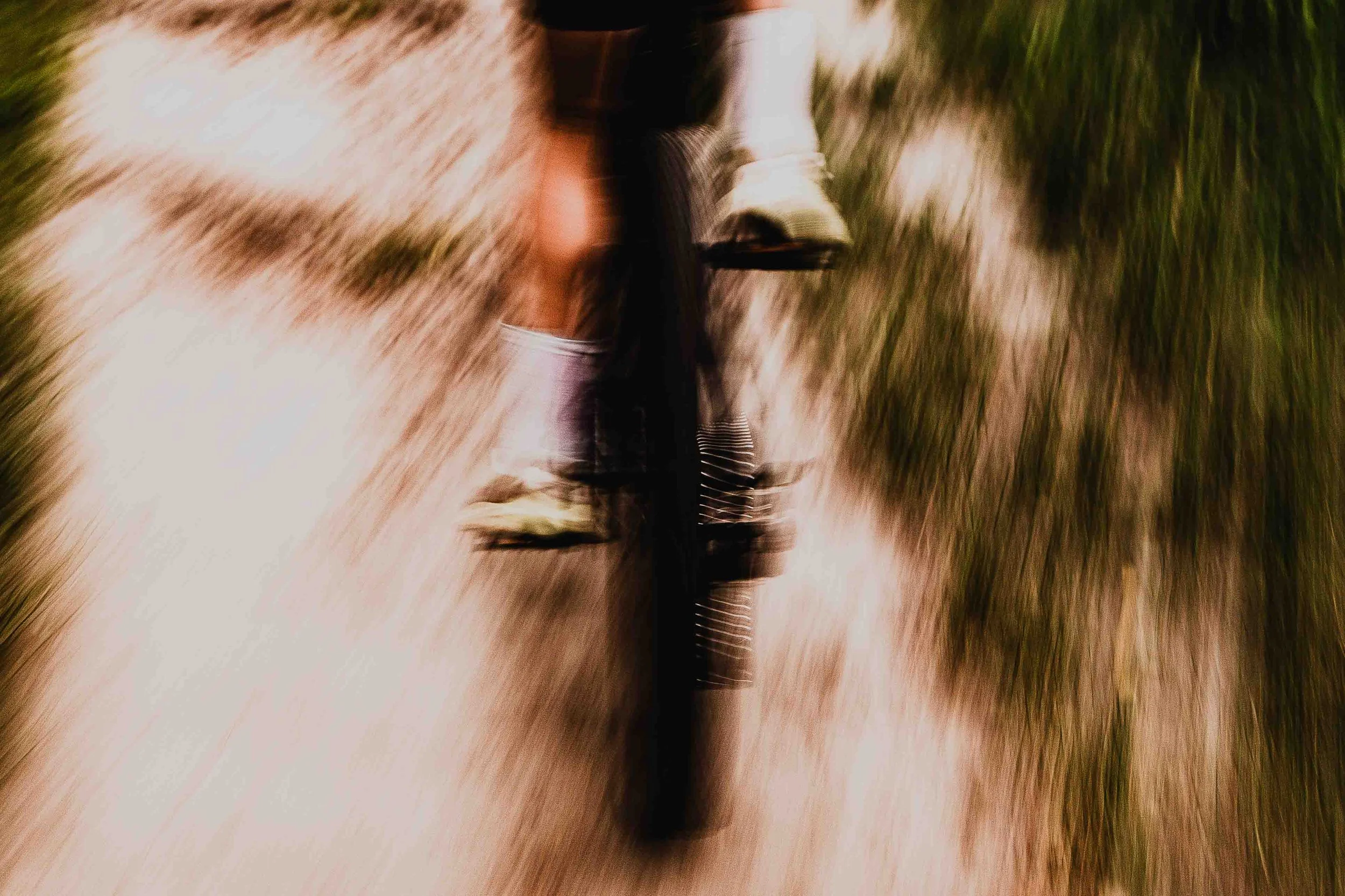 Vue en plongée d'une personne marchant dans un sentier, vue de derrière, portant un pantalon clair, des chaussures de sport et un sac à dos, avec un sol en terre et du vert en arrière-plan.