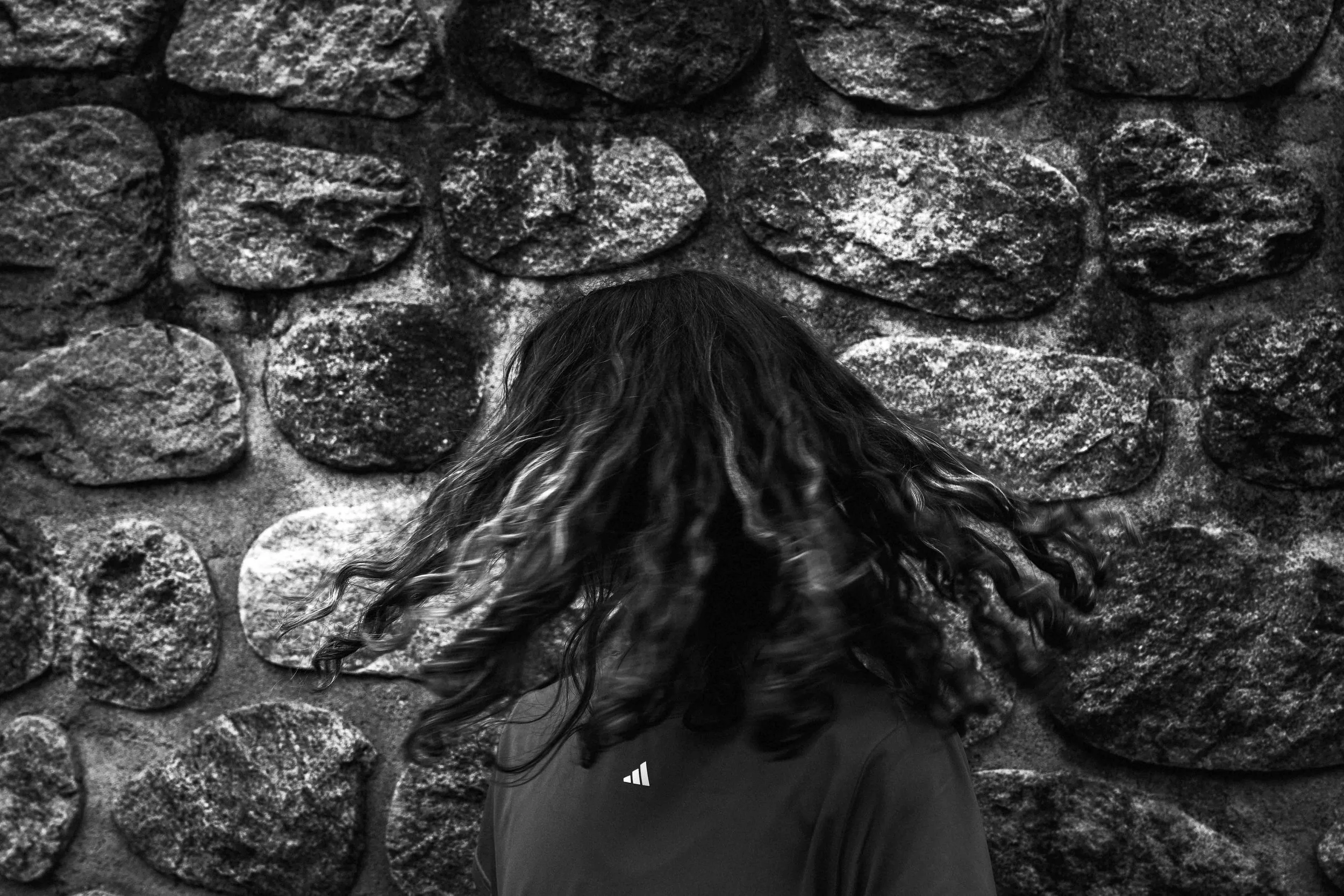 Jeune femme avec de longues cheveux bouclés se tenant devant un mur en pierre, vue de l'arrière.