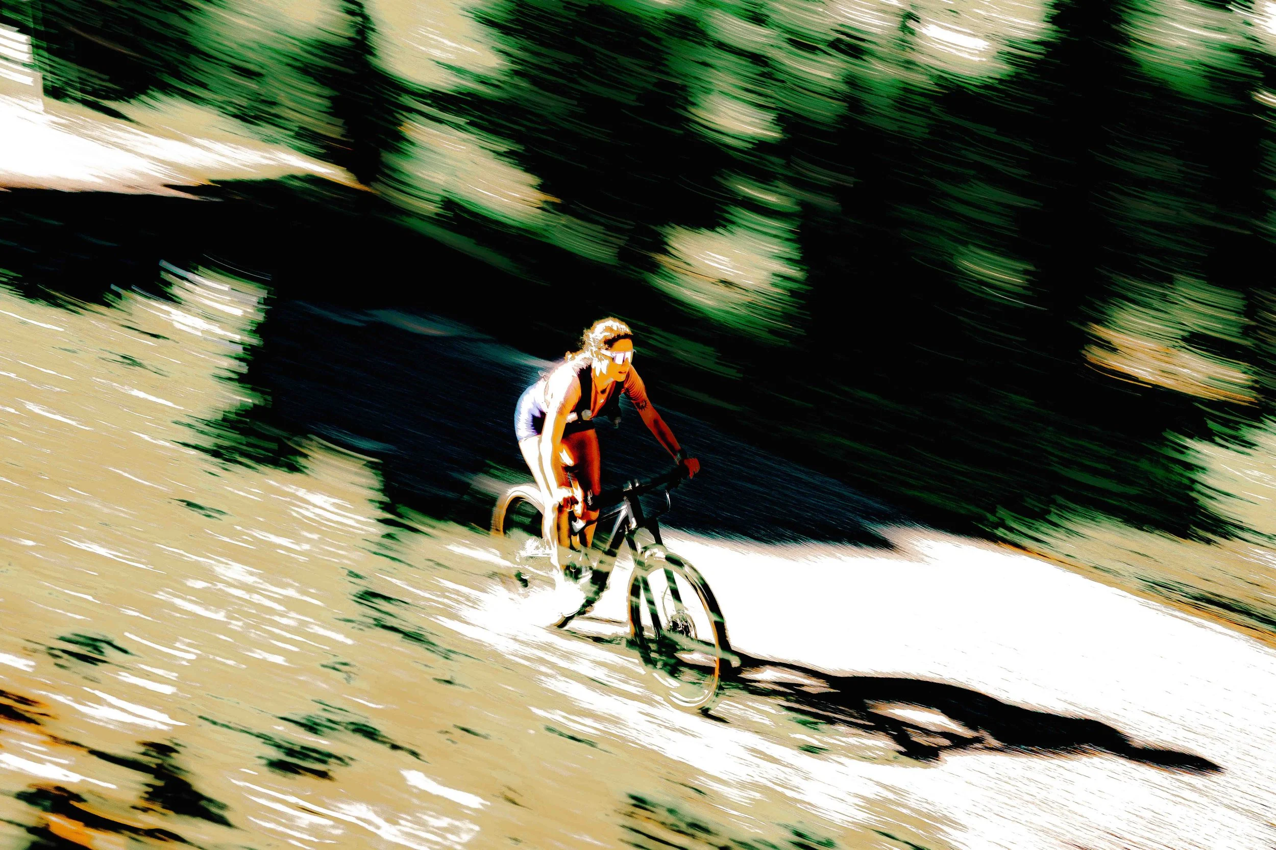 Coureur en vélo, portant des lunettes de soleil et un maillot blanc, pédalant à toute vitesse sur un sentier forestier entouré d'arbres verts.