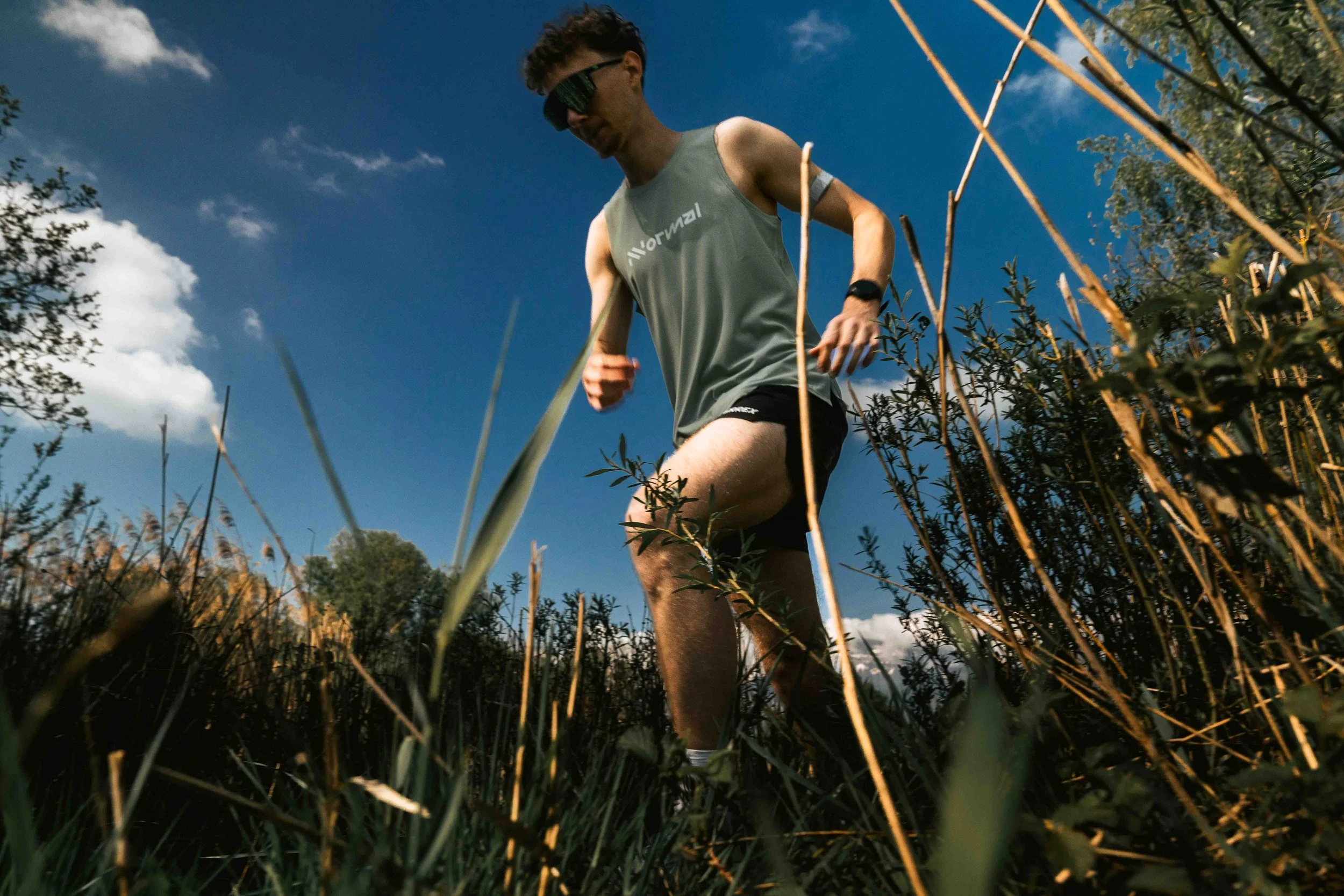 Un homme court dans un champ avec un ciel bleu et quelques nuages, portant des lunettes de soleil, un t-shirt gris et un short noir, entouré de hautes herbes et de végétation.