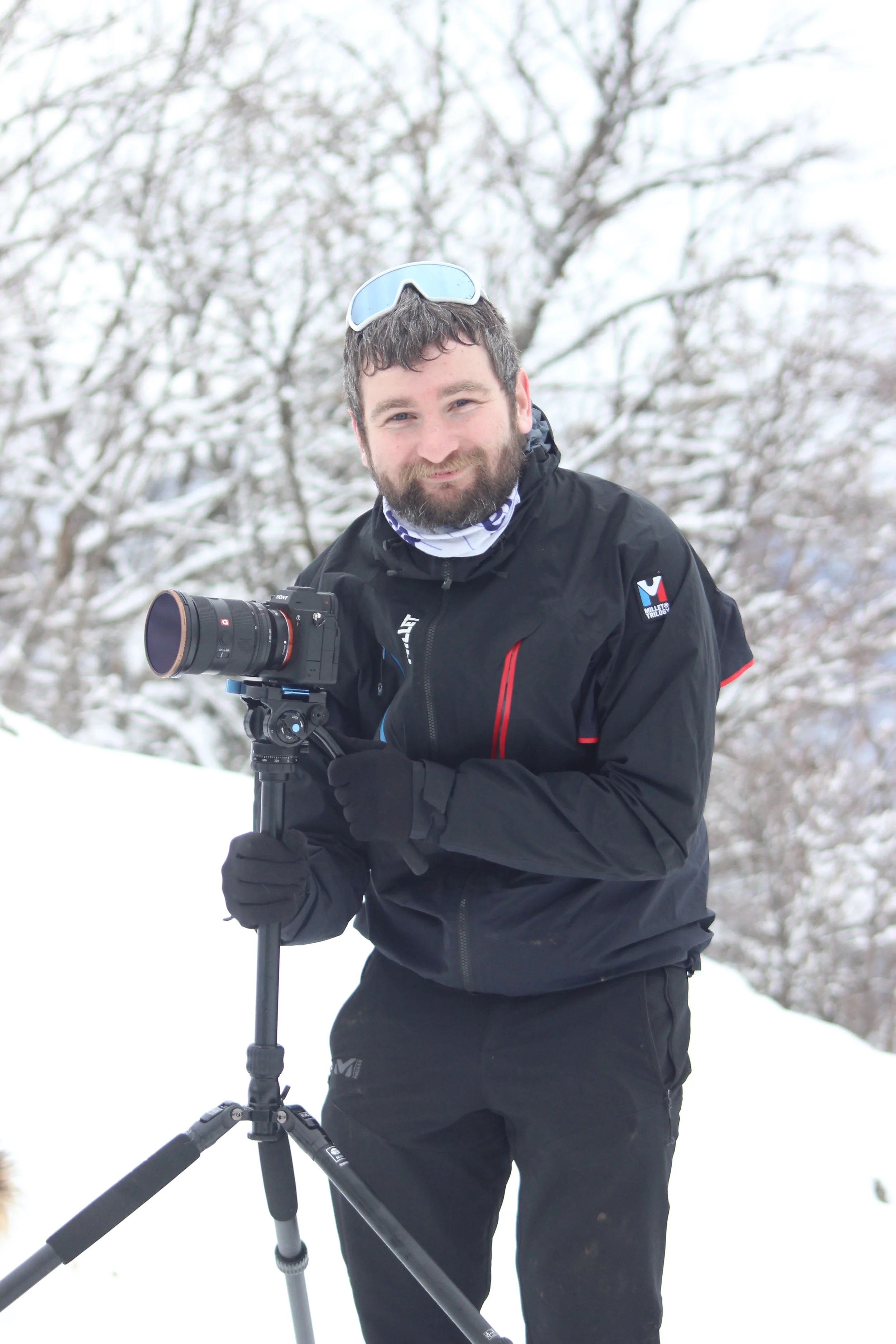 Un homme souriant dans un paysage enneigé, portant une veste noire, des lunettes de ski sur la tête, tenant un appareil photo sur un trépied.