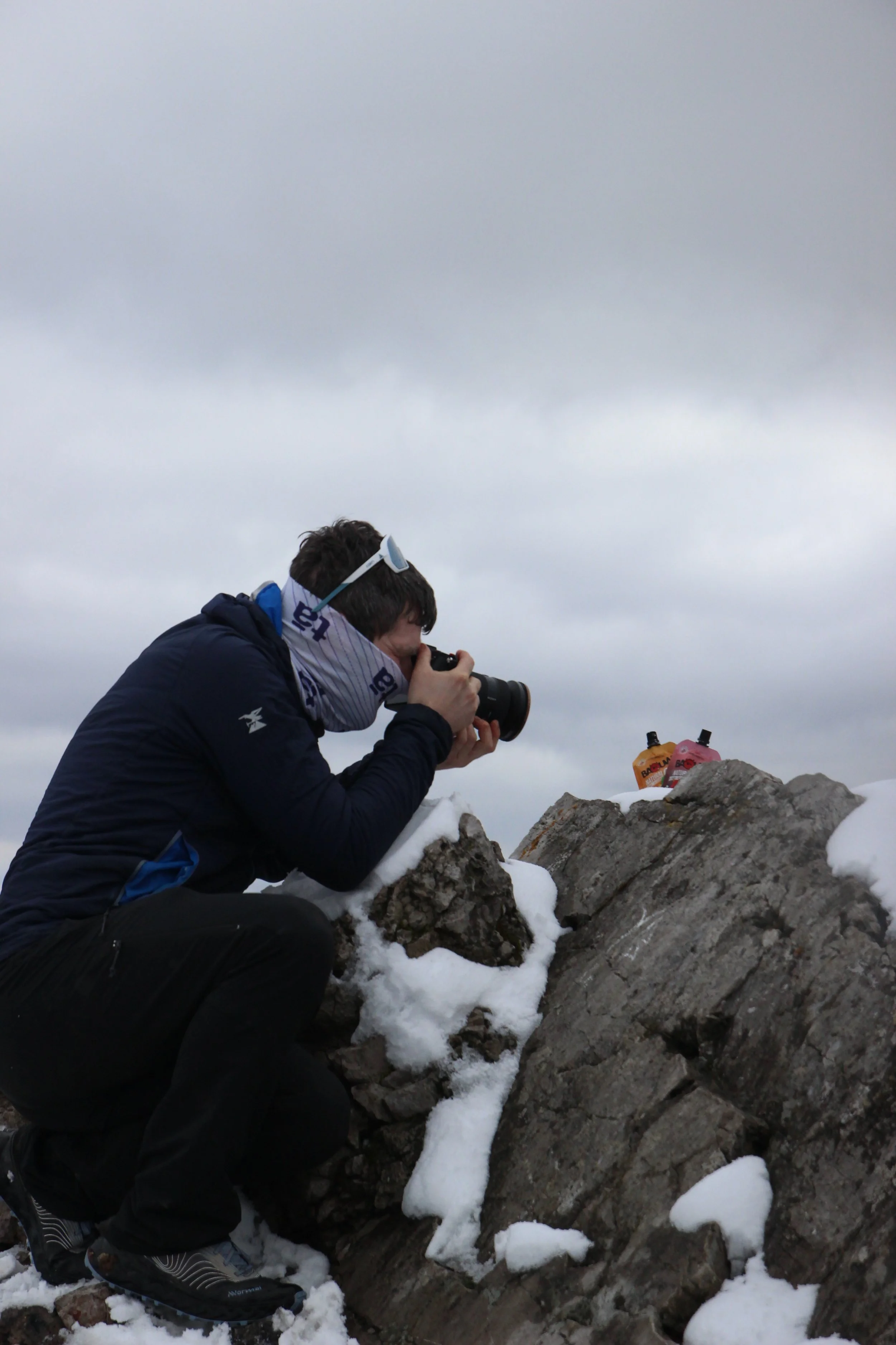 Une personne couronnée de neige prend des photos avec un appareil photo en montagne, vêtu d'une veste et de chaussures de randonnée, avec deux sacs en avant-plan et une roche enneigée sur un ciel nuageux.