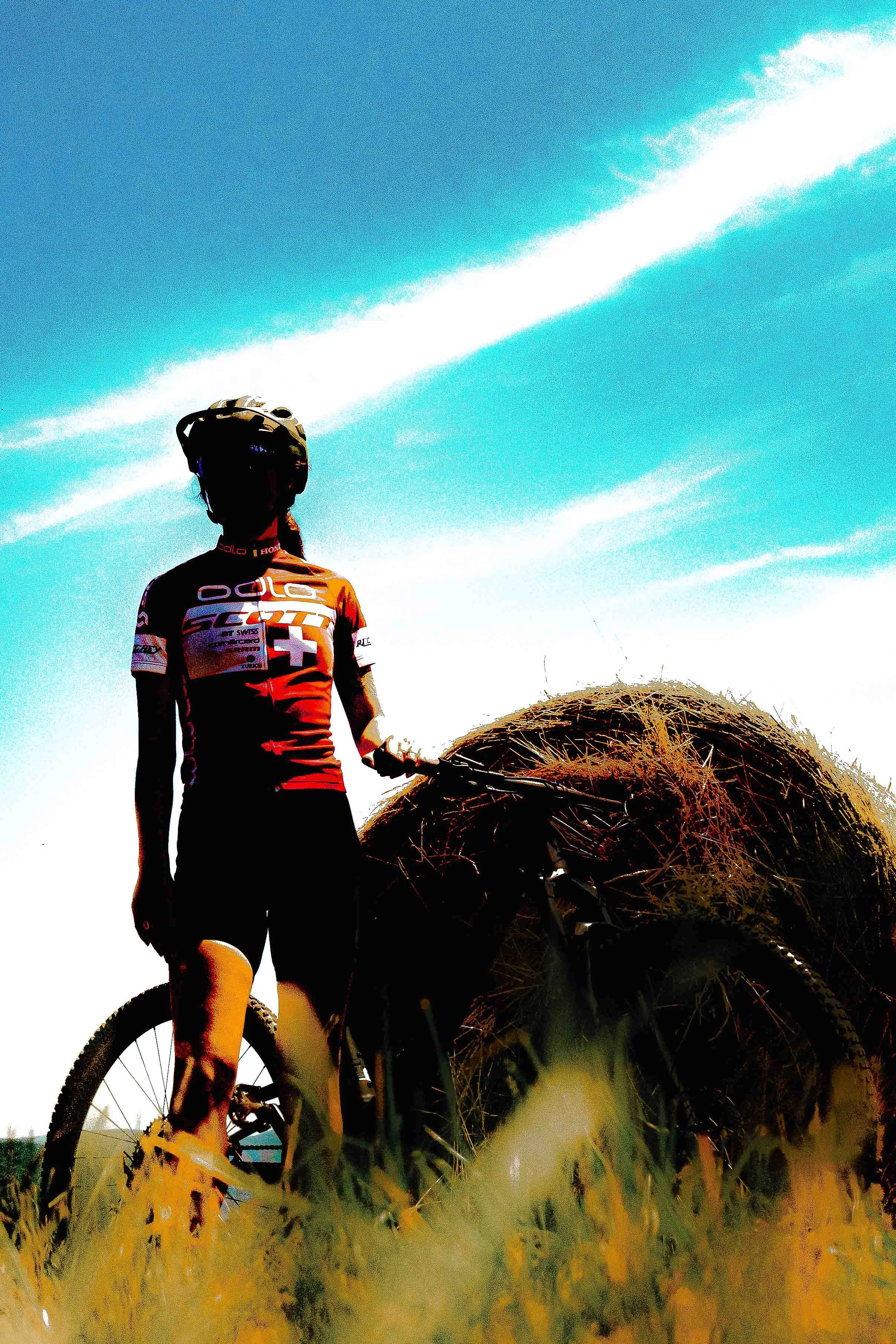 Un cycliste en tenue de sport avec un casque, debout à côté d'un vélo, près d'une récolte de foin sous un ciel bleu clair.