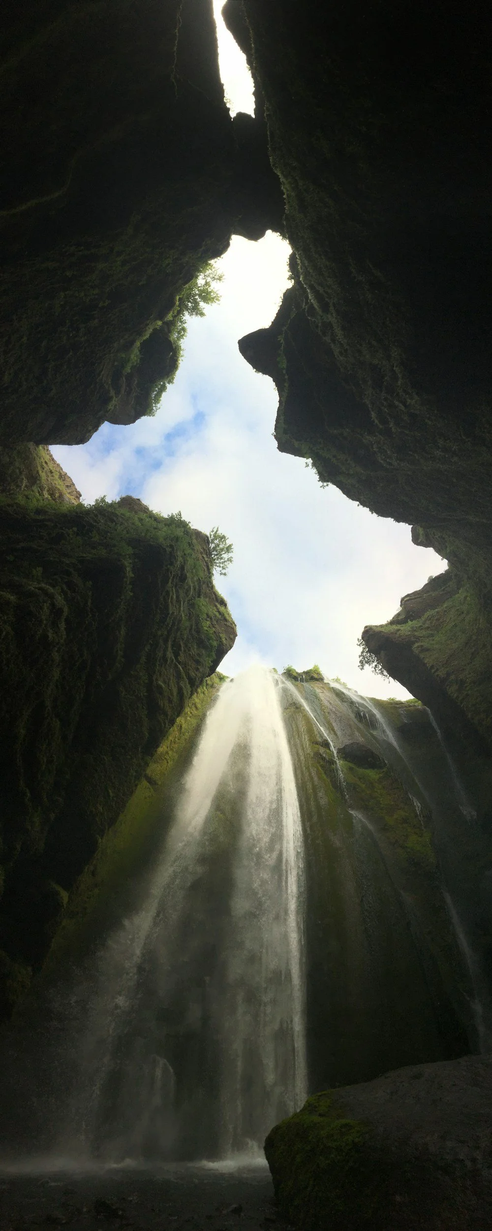 A waterfall flowing down through a narrow, moss-covered canyon with a sky view at the top.