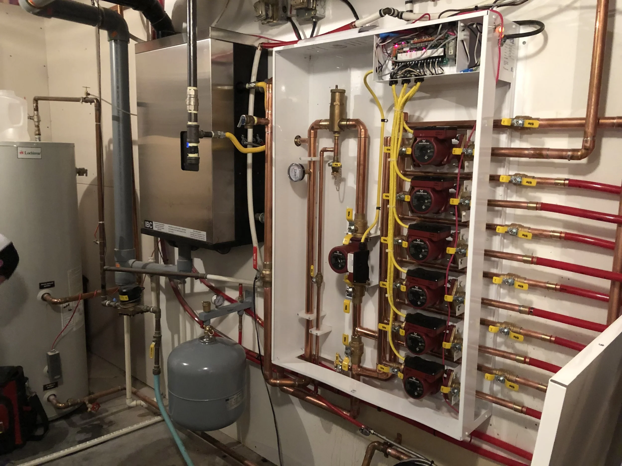 Indoor HVAC hydronic heating system with copper piping, pumps, and control components installed on a white wall.
