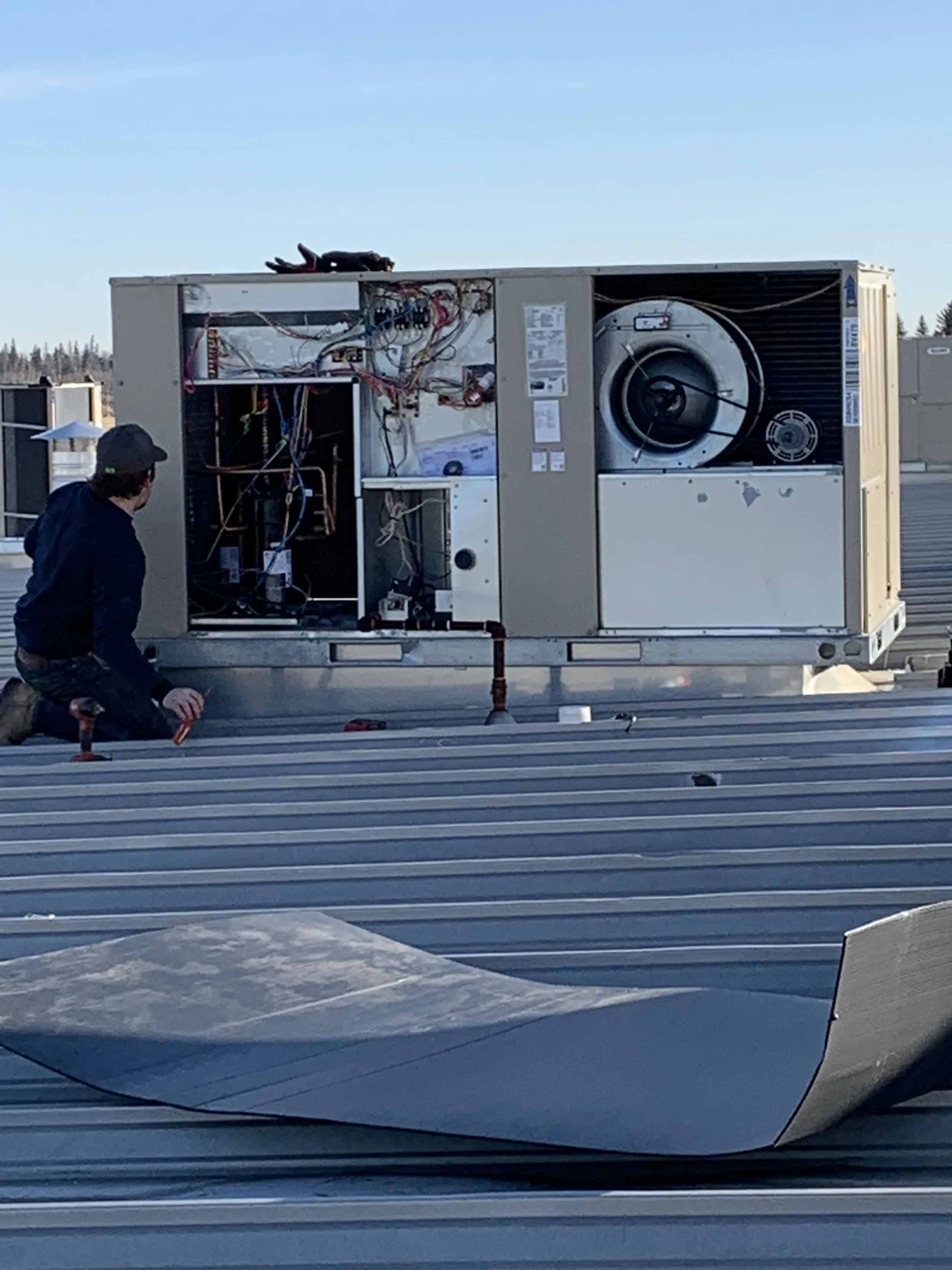 A technician kneeling on a metal rooftop working on the exposed interior of an HVAC unit, with wires and components visible.