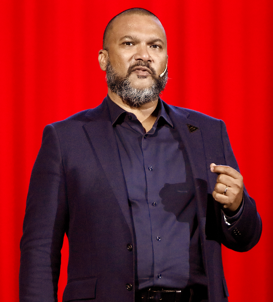 Bearded man in a dark suit speaking into a headset microphone against a red background