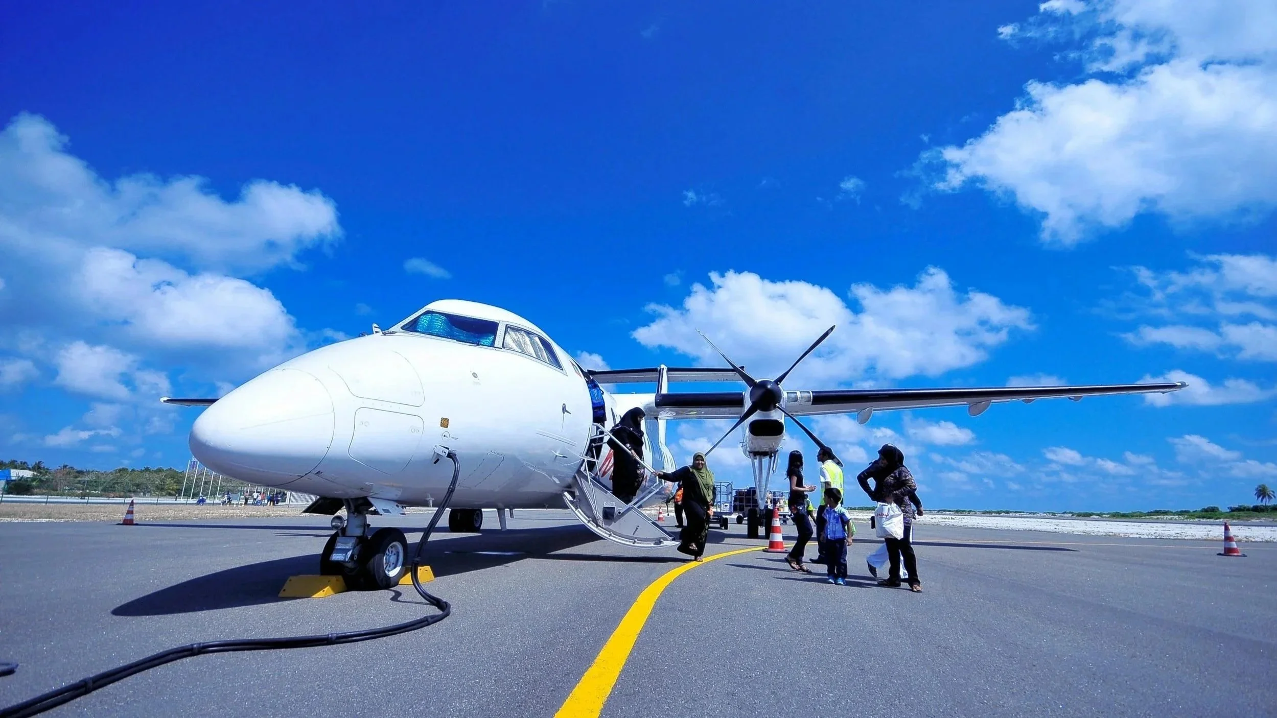 People boarding a small white propeller airplane parked on a paved runway under a blue sky with scattered clouds.