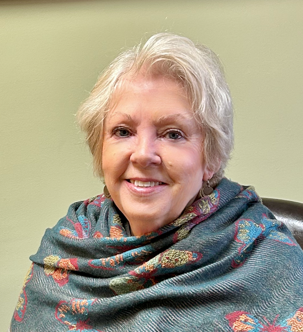 A smiling older woman with short gray hair, wearing earrings and a colorful scarf, seated against a plain light green wall.