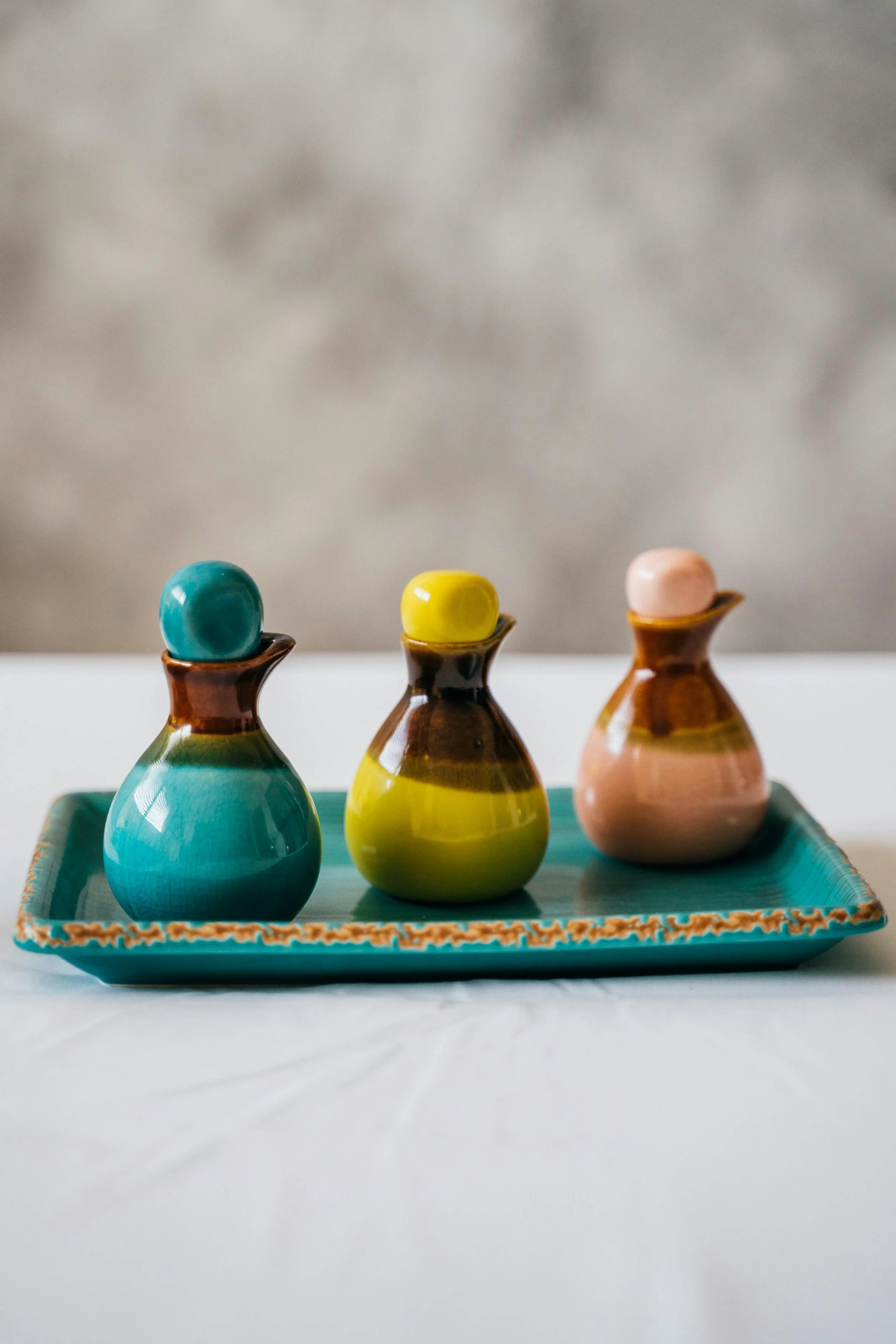Three small ceramic vases on a rectangular tray, each with a different colored ball on top, arranged on a white surface with a gray background.
