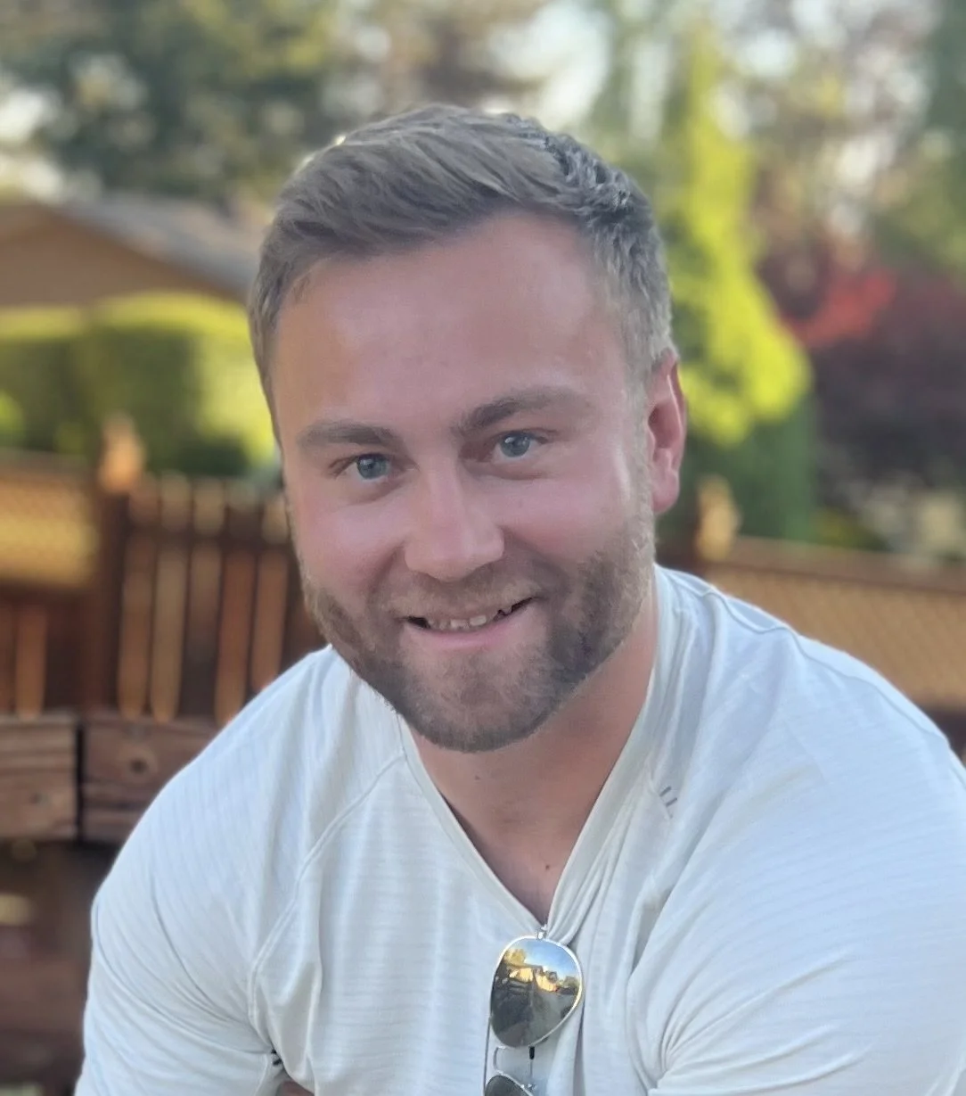A smiling man with short brown hair, a beard, and blue eyes, wearing a white shirt with sunglasses hanging from the collar, outdoors during daytime with a background of trees and a wooden fence.