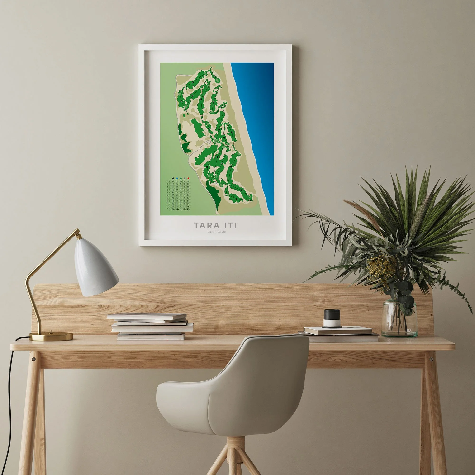 Framed map of Tara Iti Golf Club hanging on a beige wall above a wooden desk with a white chair, a desk lamp, stacks of books, and a glass vase with green plants.