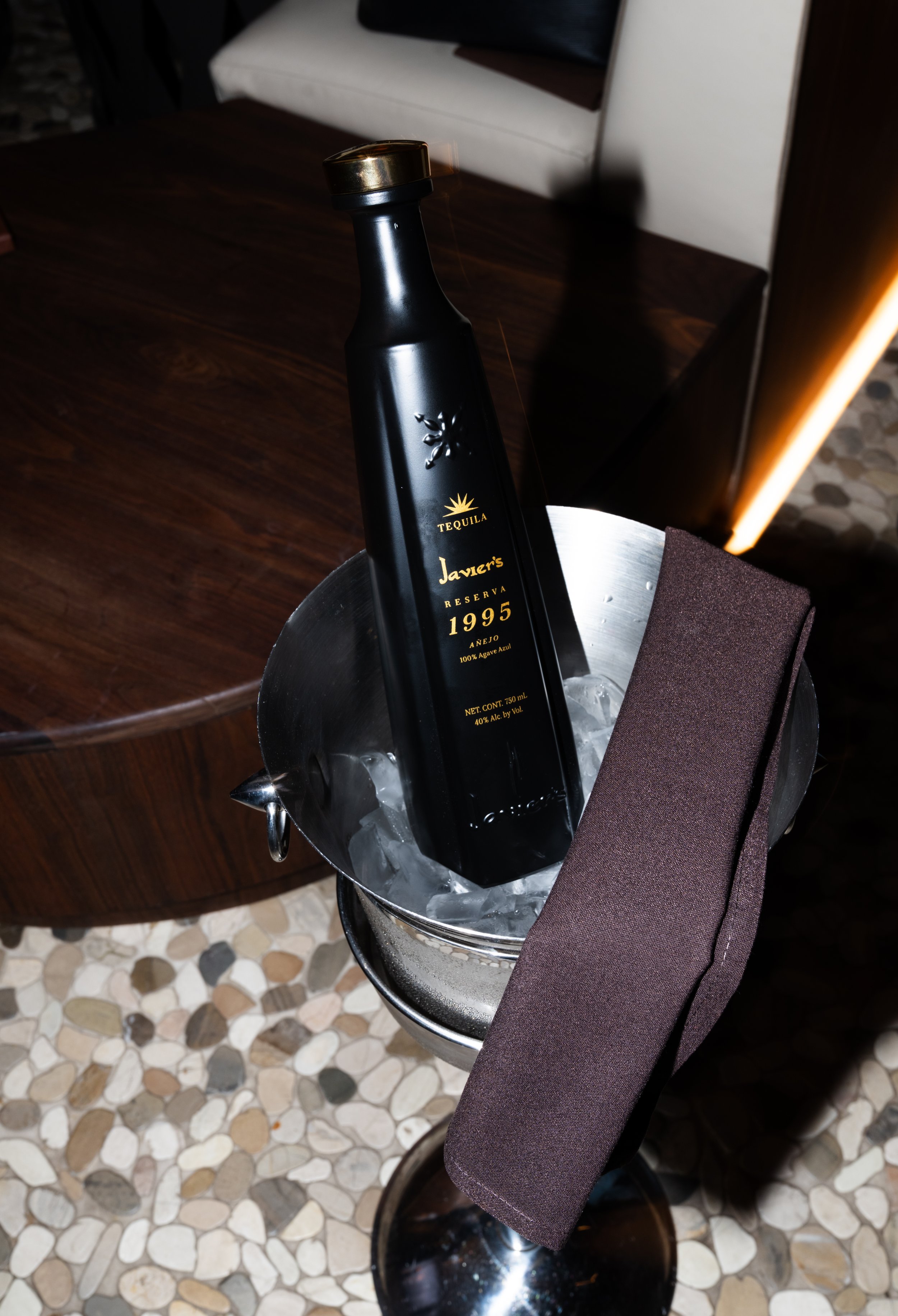 A black bottle of Javier's Reserva 1995 tequila in a silver ice bucket with ice, on a table in a restaurant.