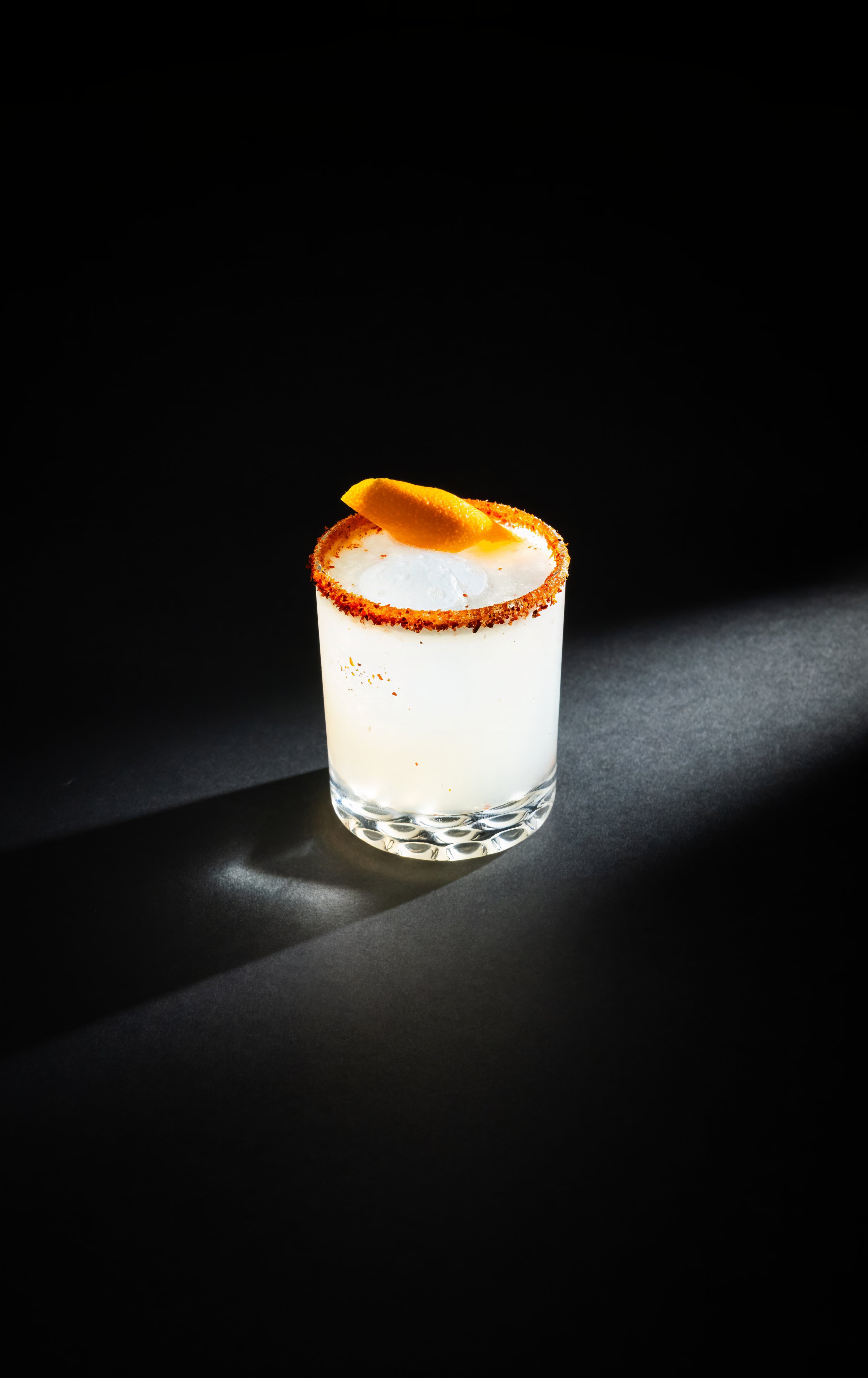A cocktail glass with a salted rim, garnished with an orange peel, filled with a margarita on a black background.