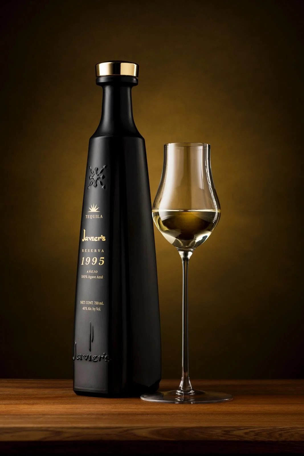 Black bottle of Javer's tequila bottle with gold accents and a filled glass of tequila on a wooden surface, against a dark brown background.