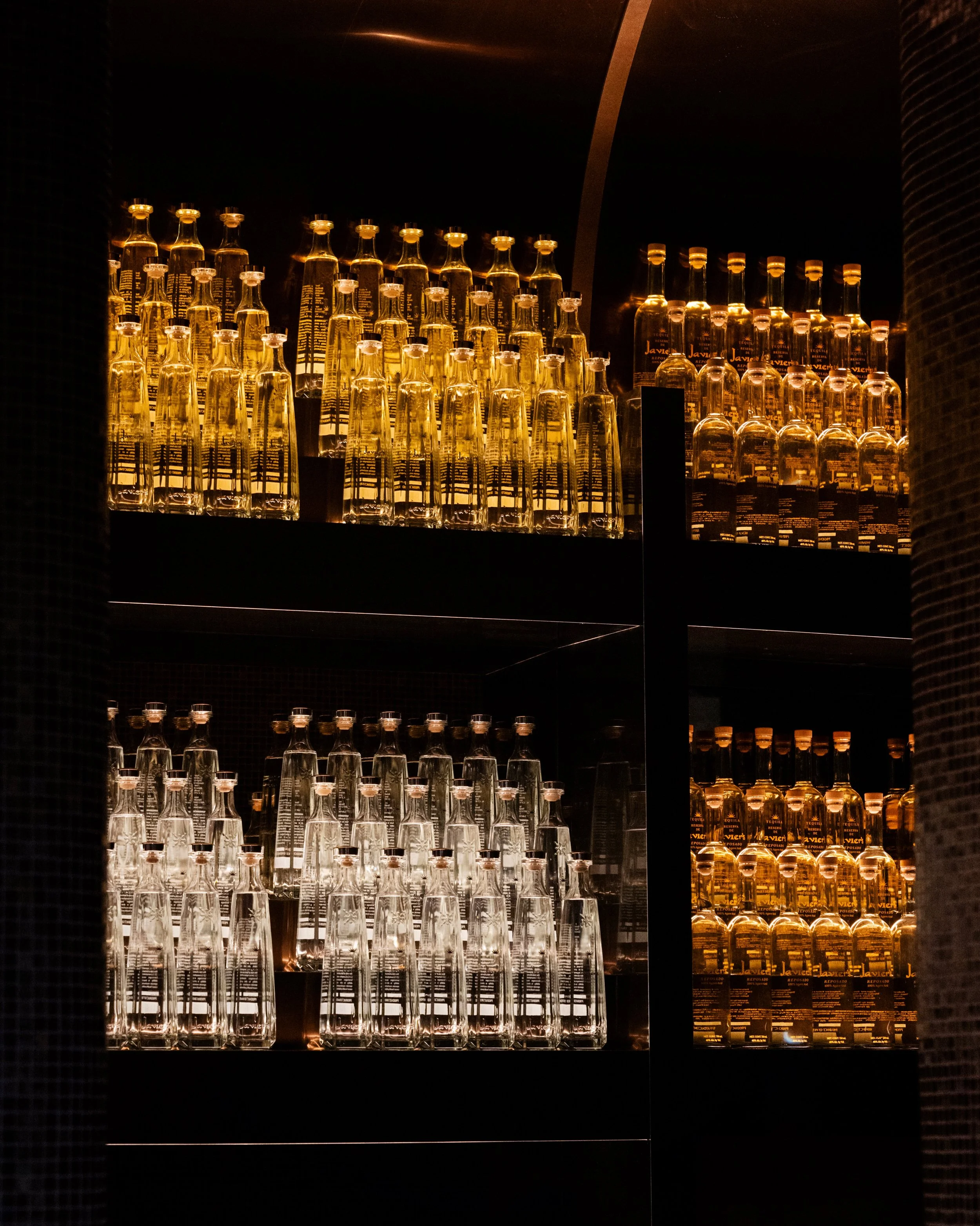 A display of clear and amber glass bottles, arranged on black shelves with warm lighting, in a bar or restaurant setting.