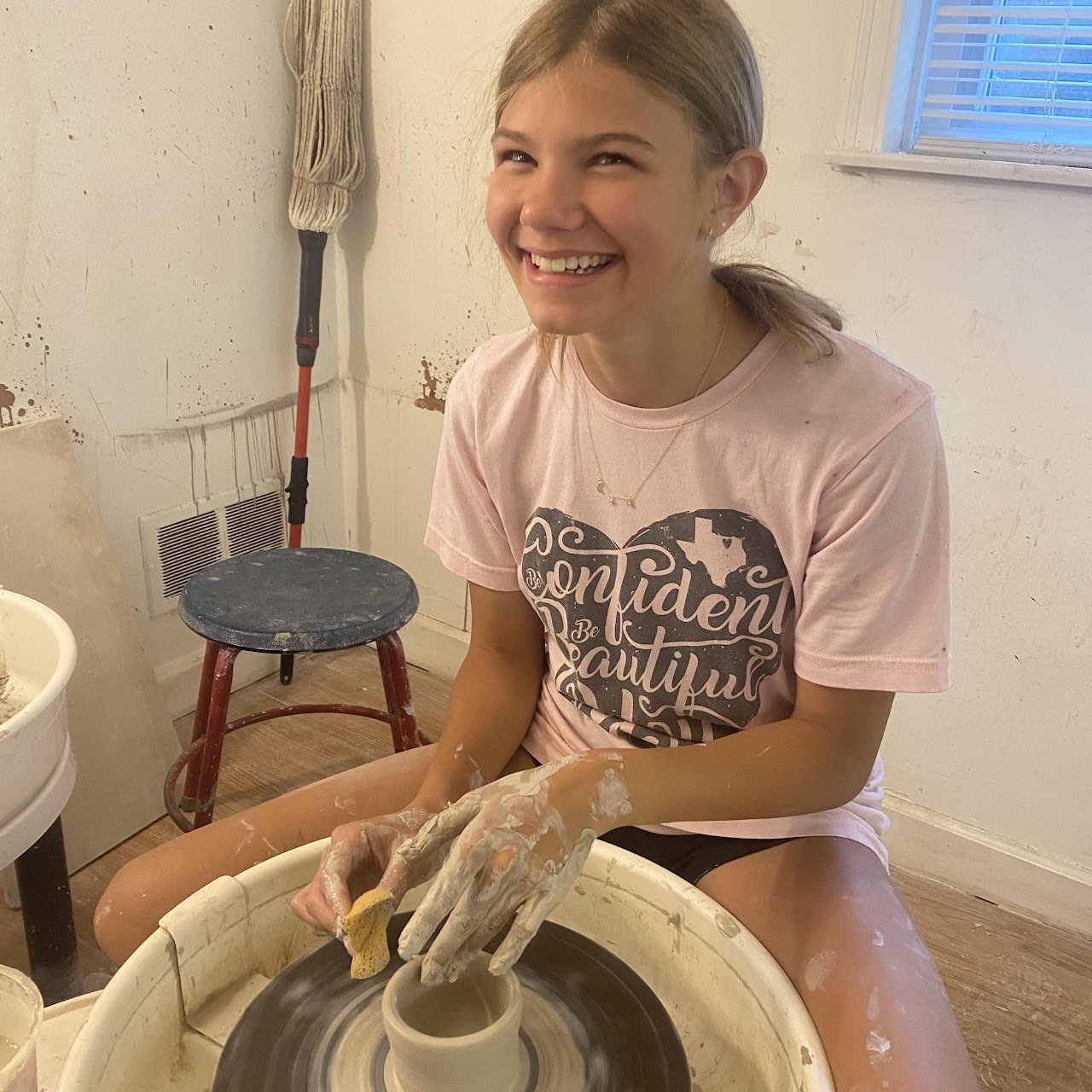 A young woman with blonde hair, smiling, and wearing a pink t-shirt, is pottery-making on a pottery wheel in a pottery studio. She has clay-covered hands and is holding a small sponge and shaping a ceramic cup.