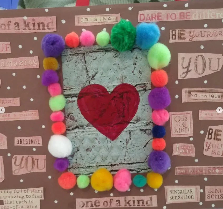 Decorative collage featuring a centered red heart painted on weathered wood, surrounded by multicolored pom-poms, with cut-out phrases and words on a brown background that emphasize uniqueness and kindness.