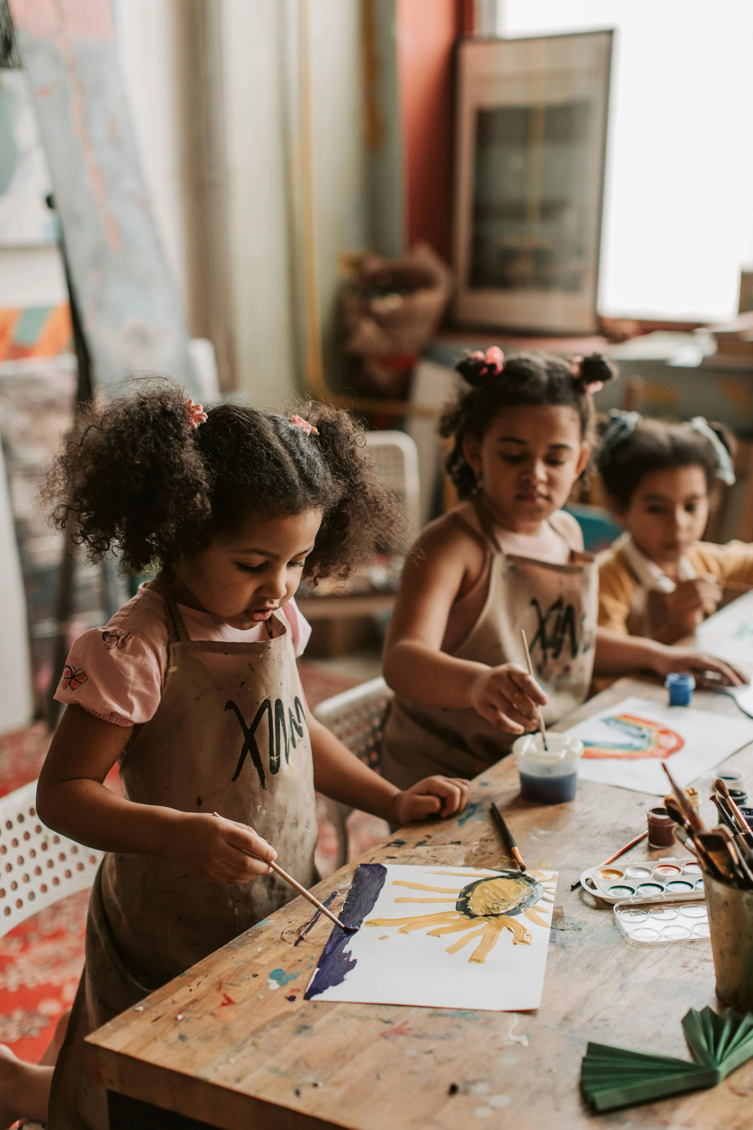 Three young girls with curly hair and aprons painting at a table in an art classroom.