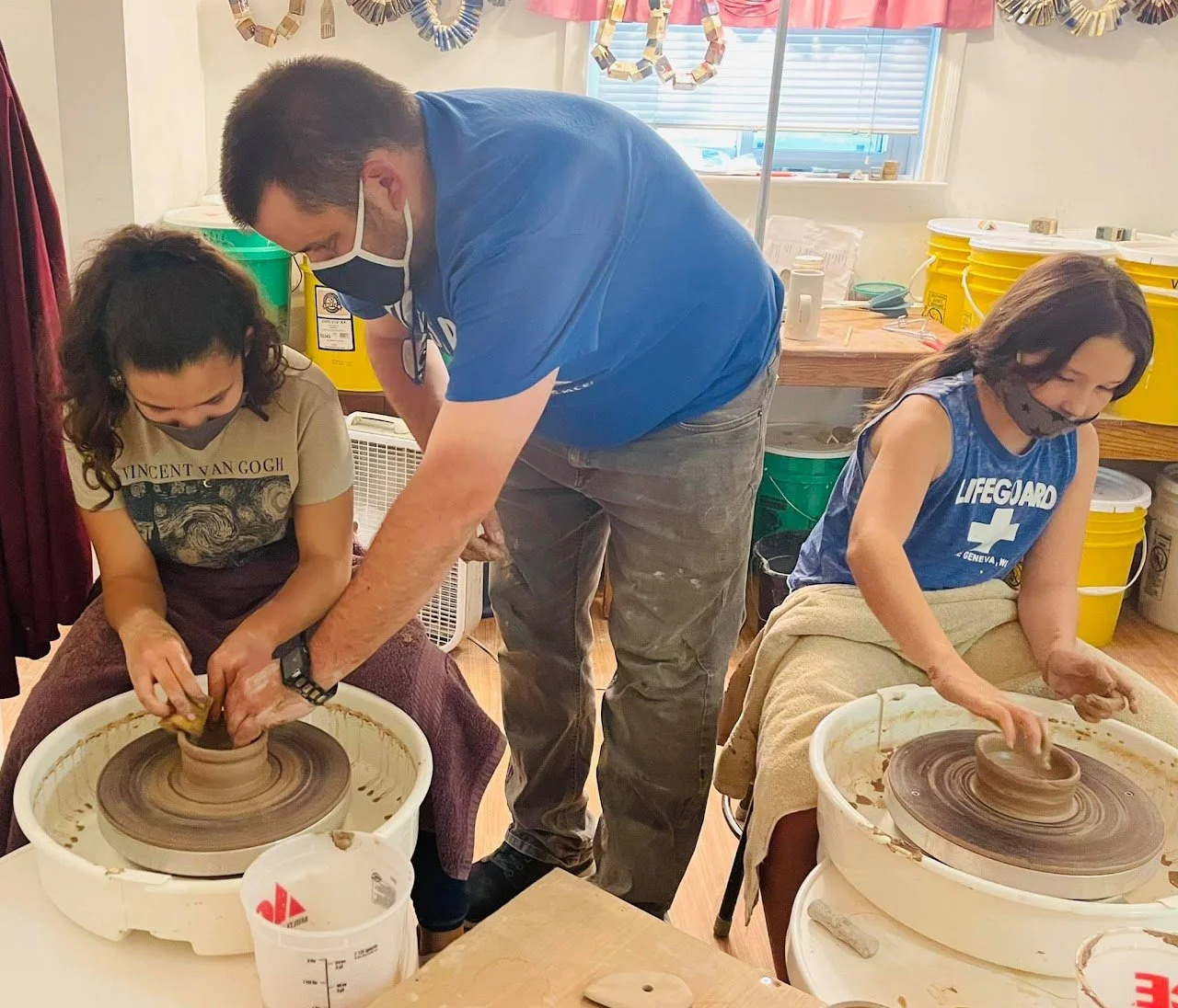 A man with two young girls using pottery wheels to shape clay in a classroom setting.