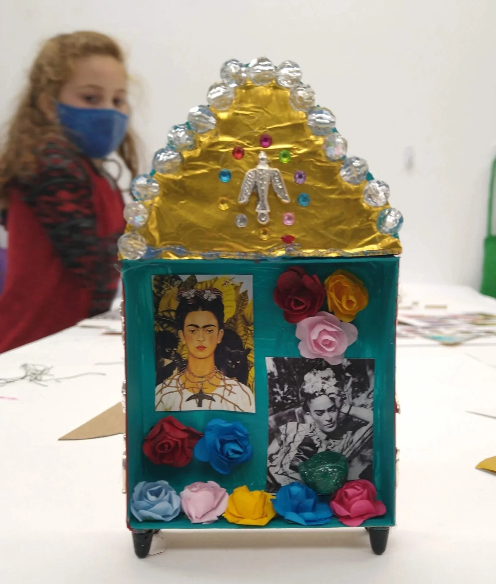 A colorful, decorated display with Frida Kahlo images, paper flowers, shiny ornaments, and a gold top, with a girl wearing a face mask in the background.