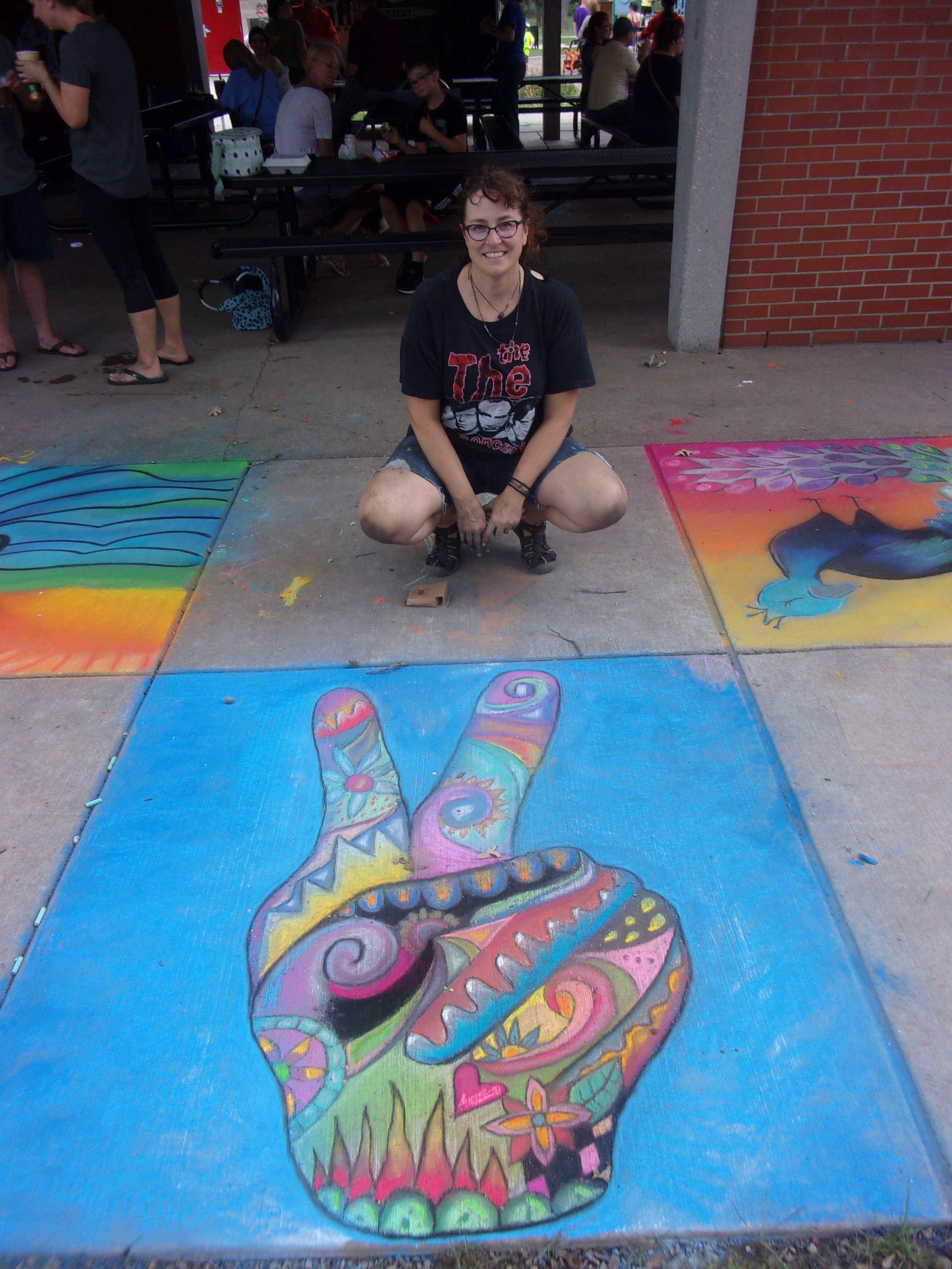 A woman squatting on the sidewalk in front of colorful chalk art depicting a skull and a peace sign made with vibrant, intricate patterns. People are sitting at picnic tables in the background.