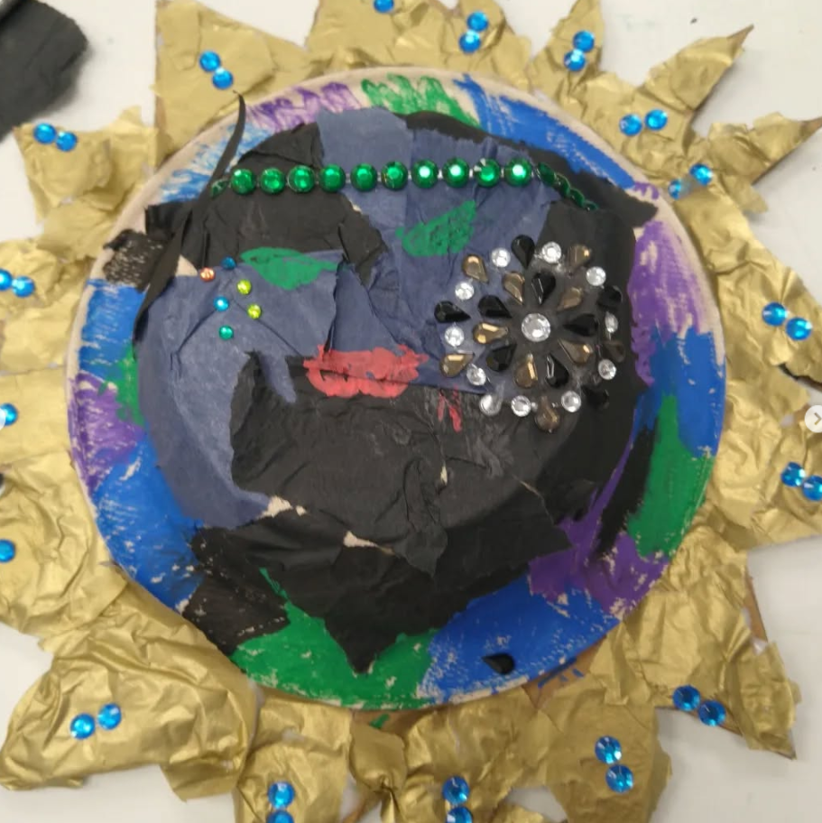 A colorful handmade art piece featuring a portrait of a woman with dark hair and green eyes, decorated with rhinestones, surrounded by crumpled gold paper and blue beads.