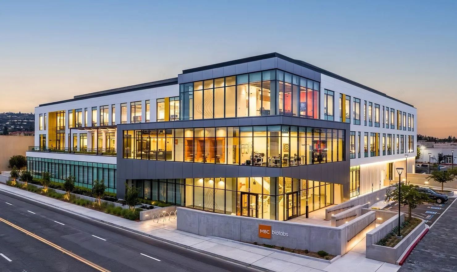 Modern office building at dusk with large glass windows, landscaping, parking lot, and a sign that reads "MBC biolabs."