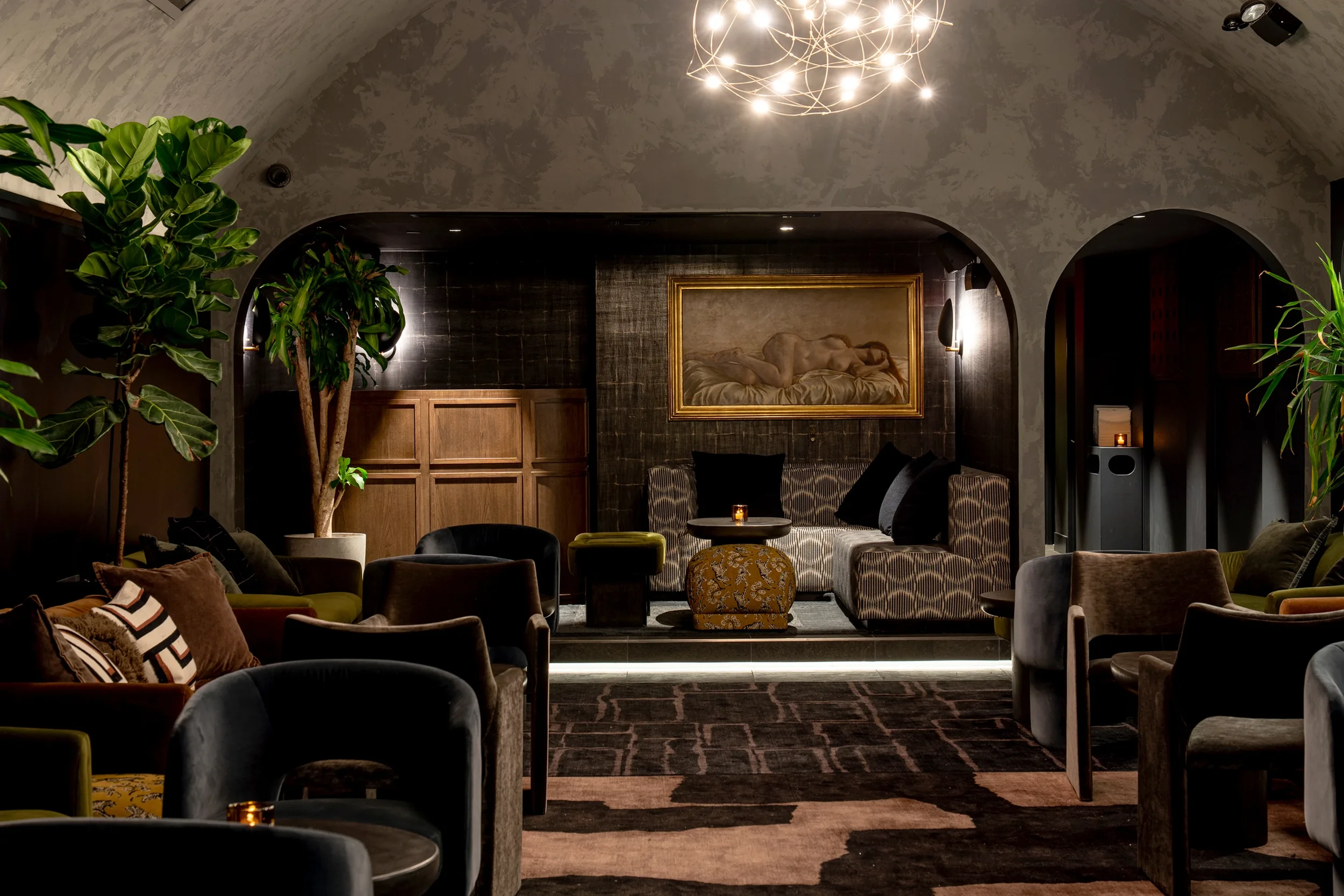 Photograph of Broome St. Project interior. A cozy, dimly lit lounge with plush chairs, patterned sofas, and large indoor plants.