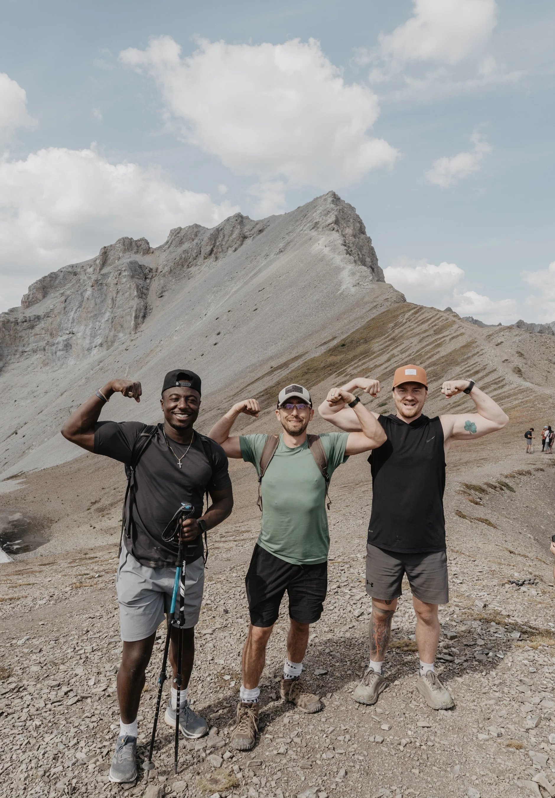 Three men standing outdoors on a rocky mountain trail, flexing their biceps and smiling at the camera with a mountain and cloudy sky in the background.