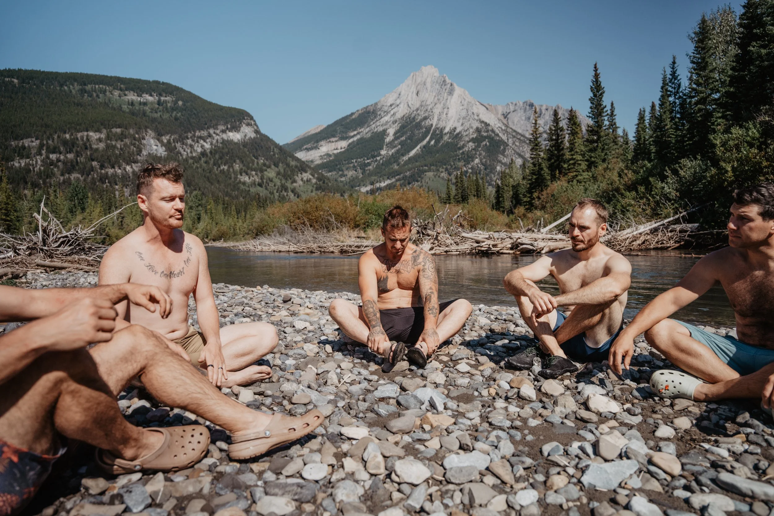 Five shirtless men sitting on a rocky riverbank, with mountains and trees in the background, during daytime.