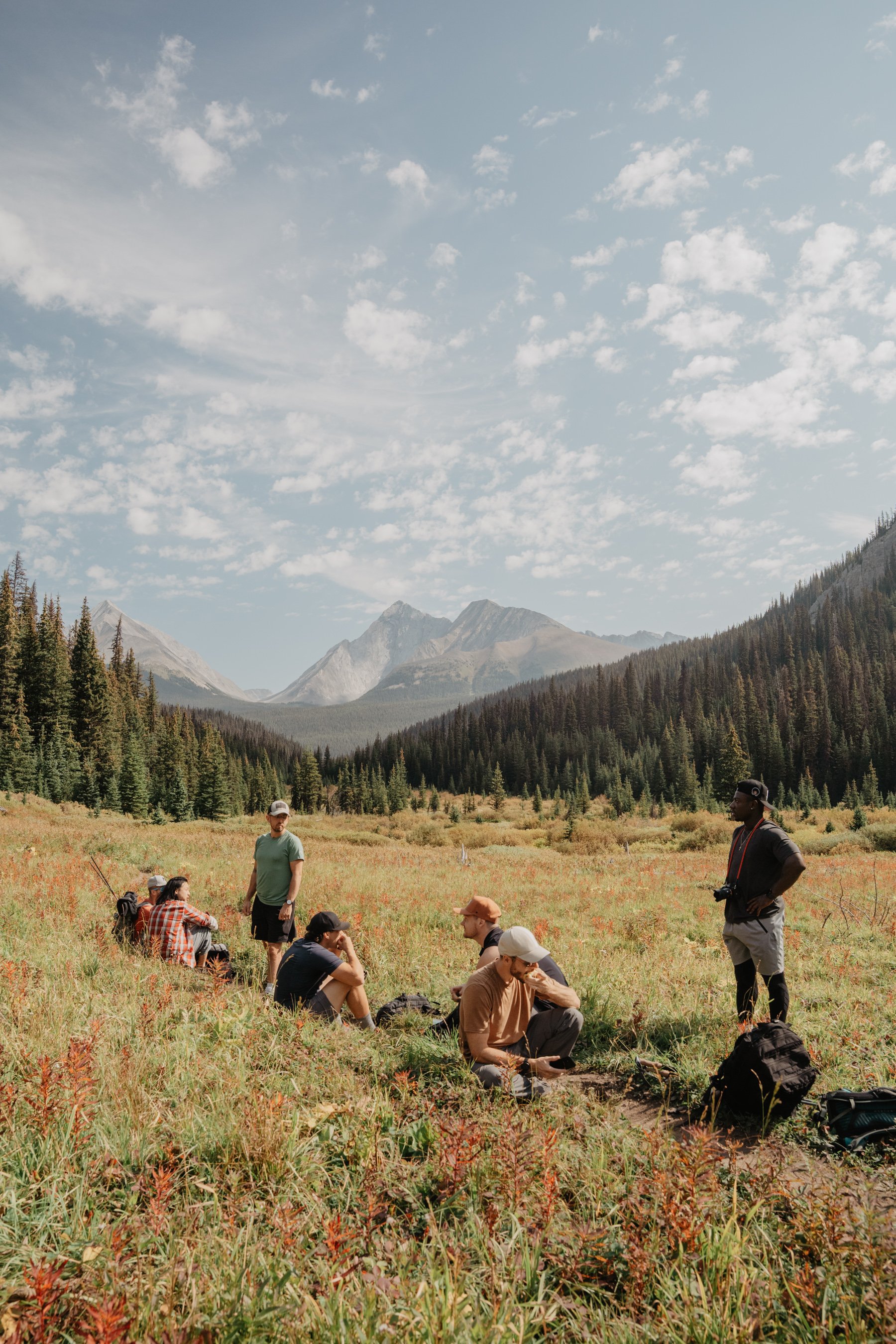 A group of people taking a break during a hike in a scenic mountain valley with tall pine trees and snow-capped peaks under a partly cloudy sky.