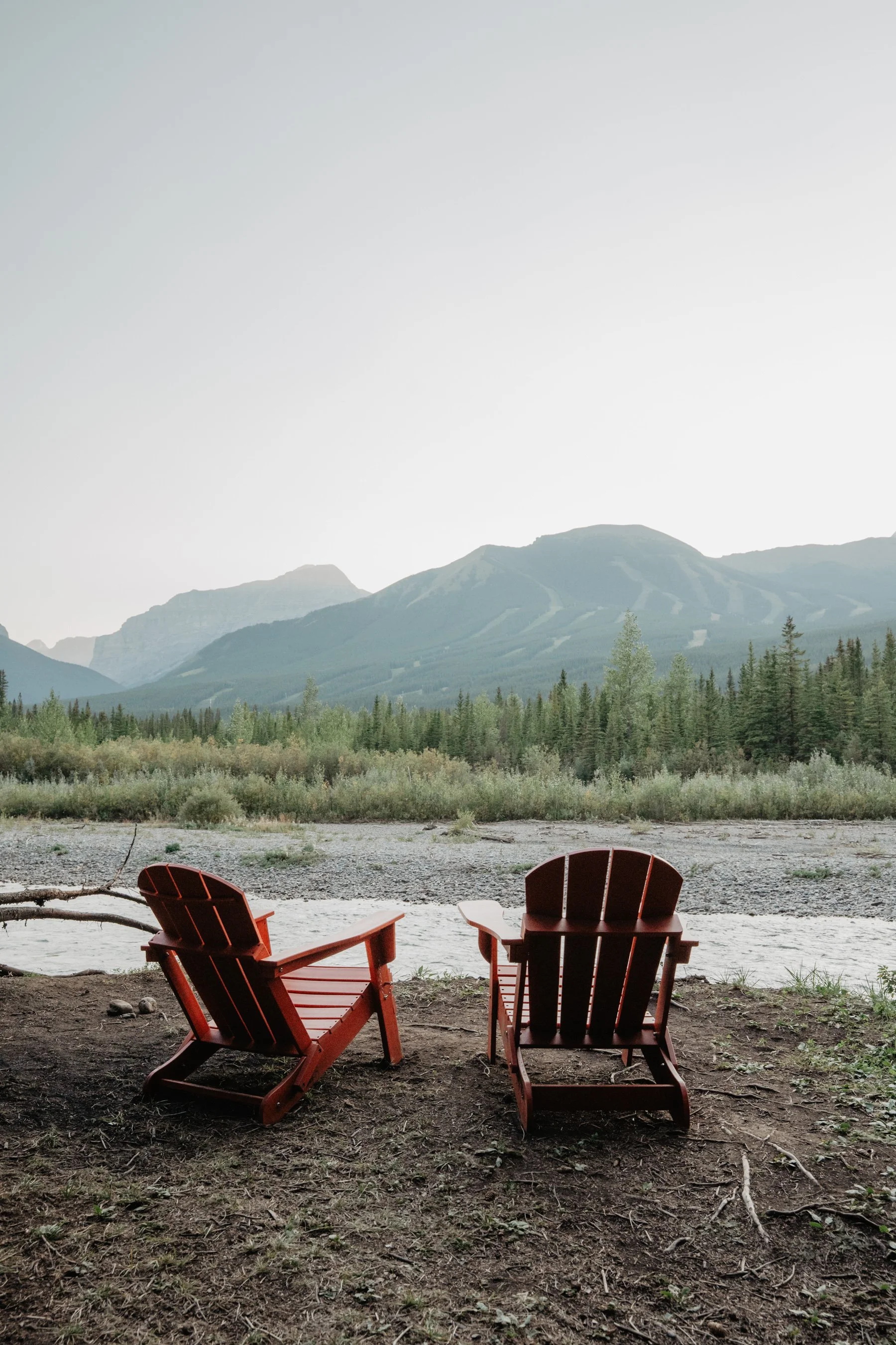 Two red Adirondack chairs on a dirt ground facing a river with lush green trees and mountains in the background under a clear sky.