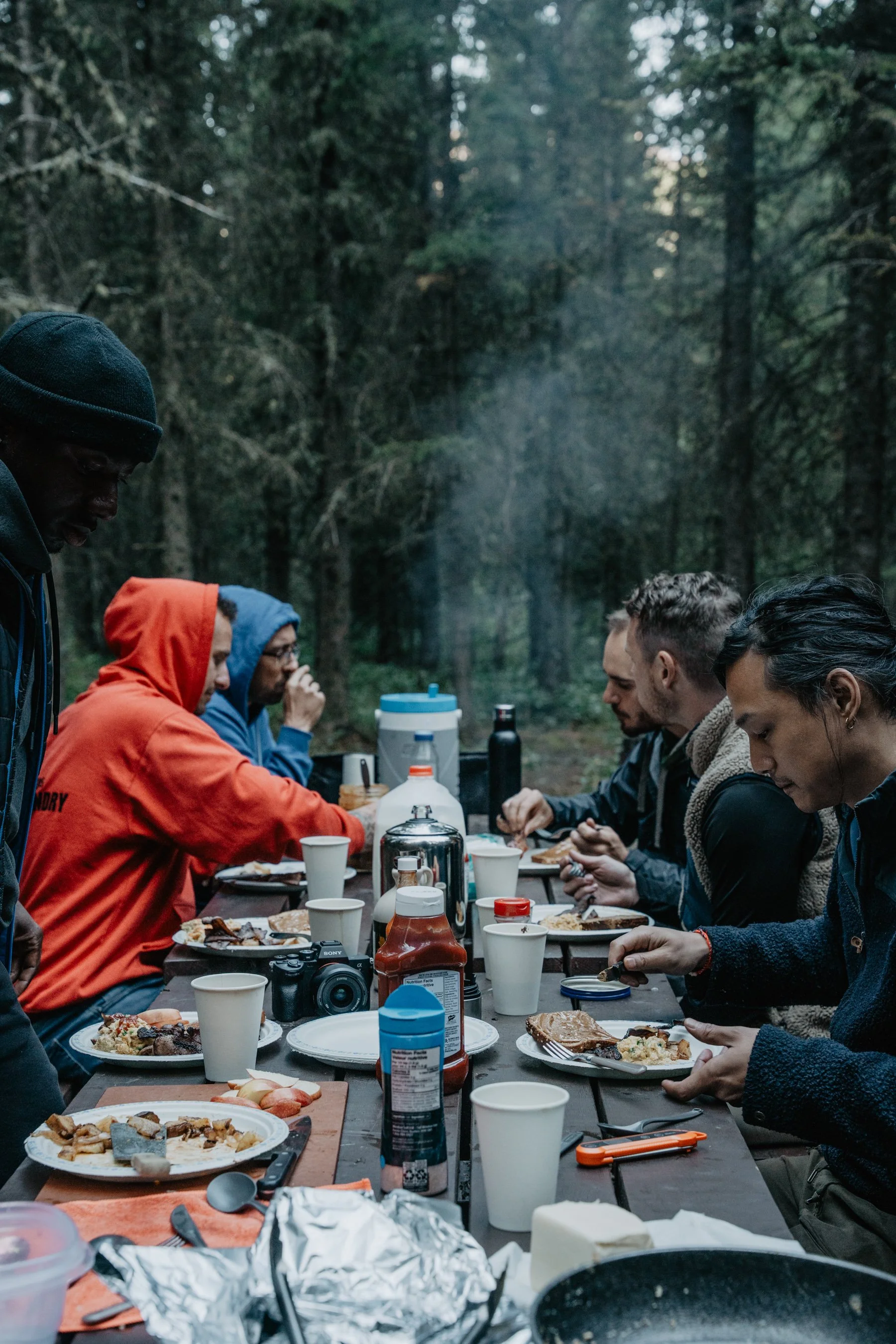 People gathered around a long outdoor table in a forest, having breakfast or brunch with plates of food, drinks, and condiments.