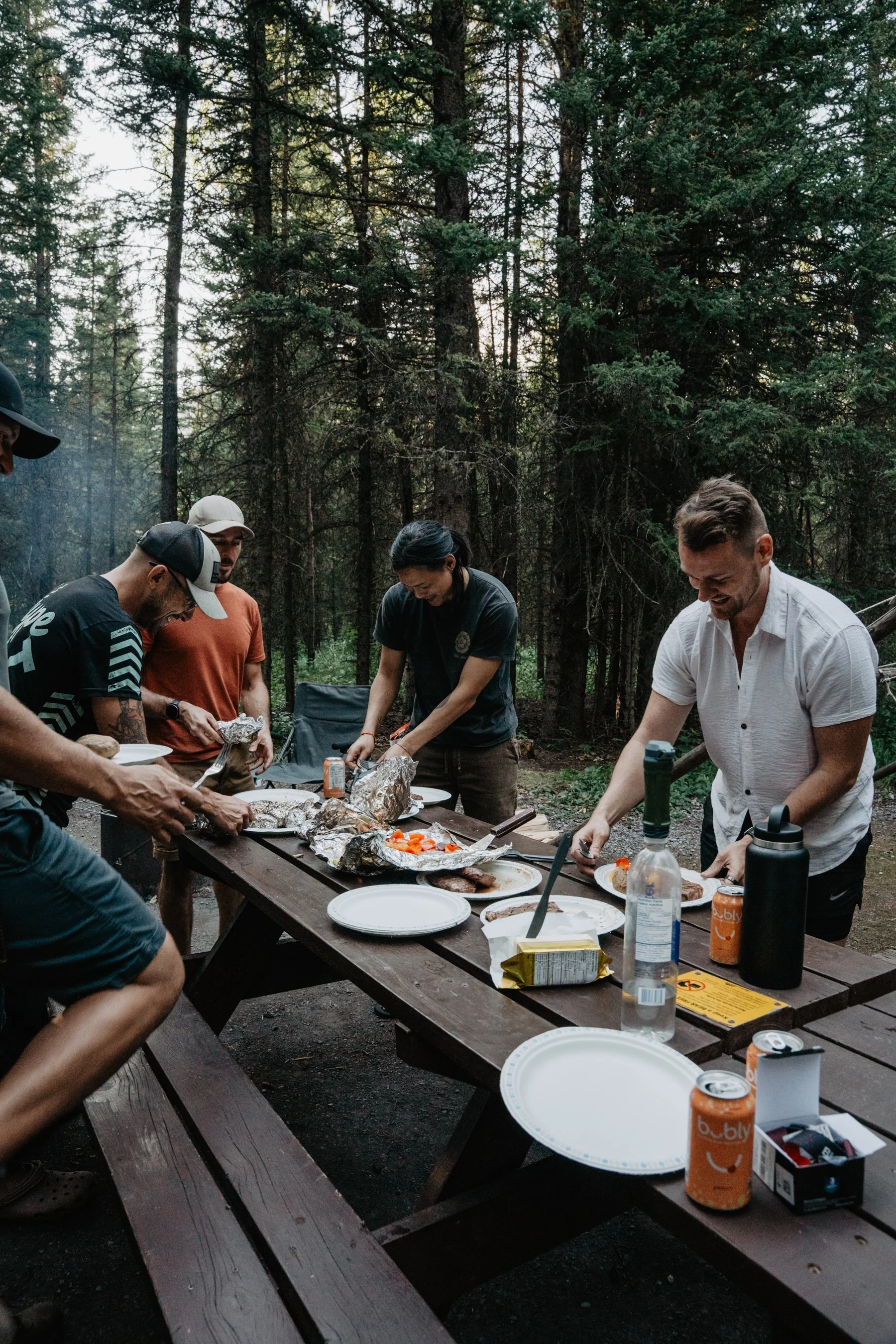 Group of people enjoying a barbecue picnic outdoors in a forest, with food and drinks on a wooden picnic table.